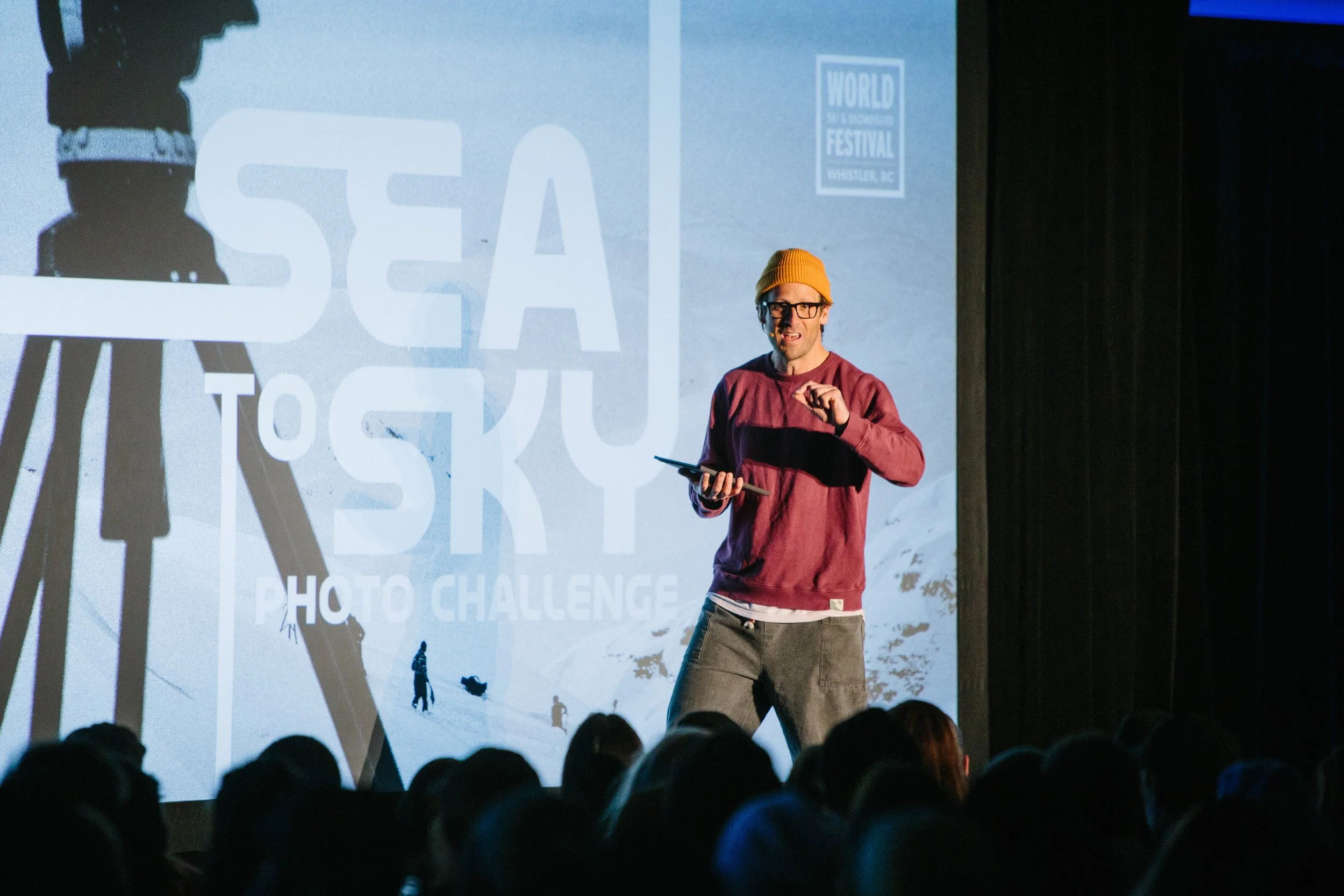 A man wearing a yellow beanie, glasses, and a maroon sweatshirt on stage giving a presentation. Behind him is a large screen with a snowy mountain and the text 'Sea to Sky Photo Challenge.' An audience is visible in the foreground.