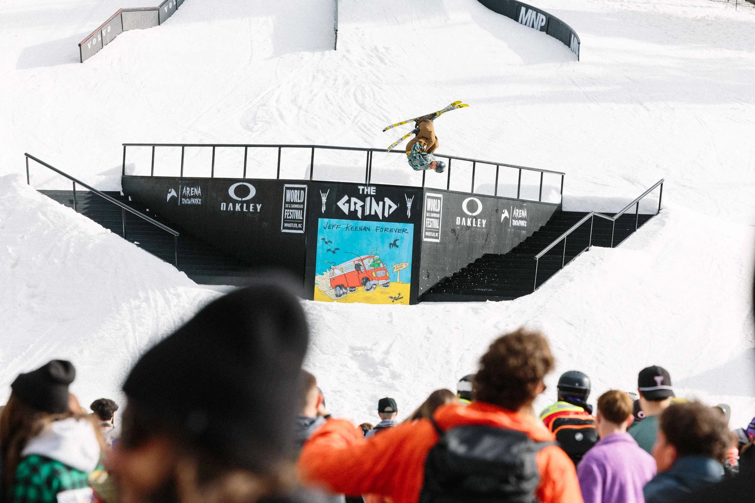 A snowboarder mid-air performing a trick at a snowboarding competition with a crowd watching below and a black ramp with signs.