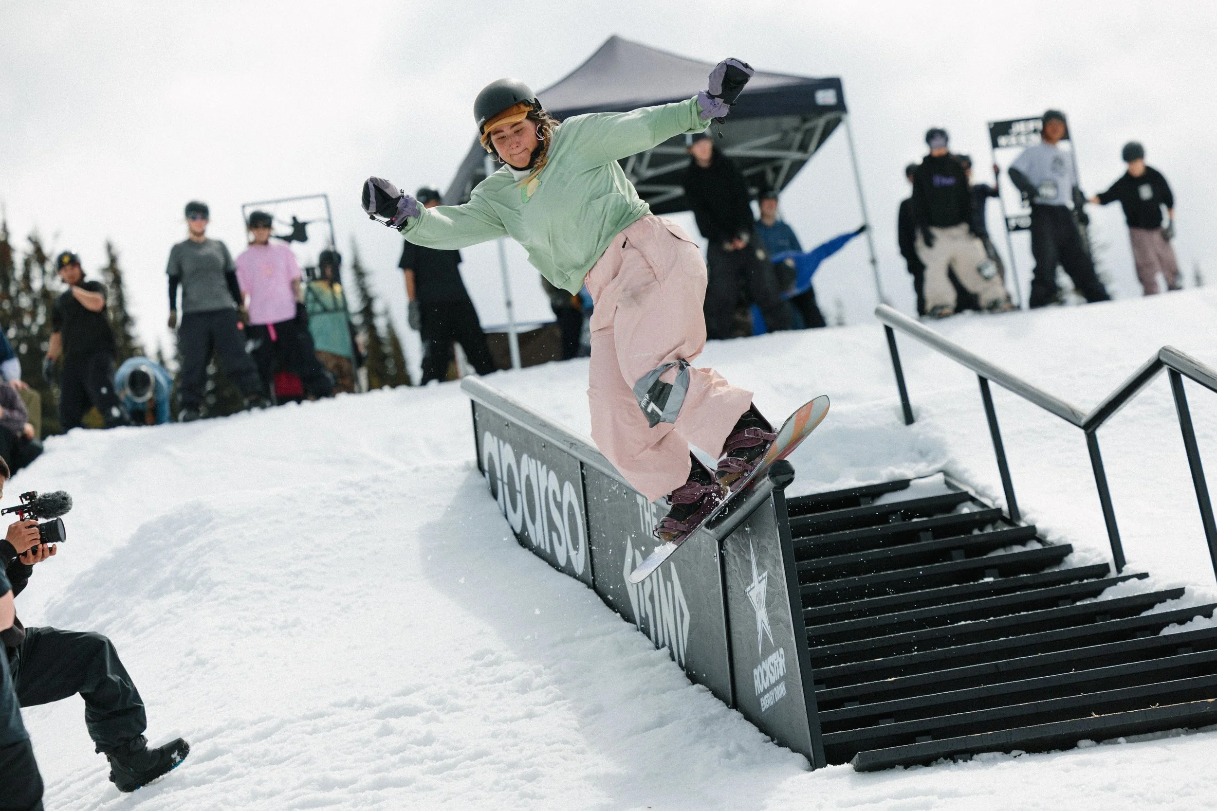 A young person snowboarding down a rail in a snow park with onlookers and photographers in the background.