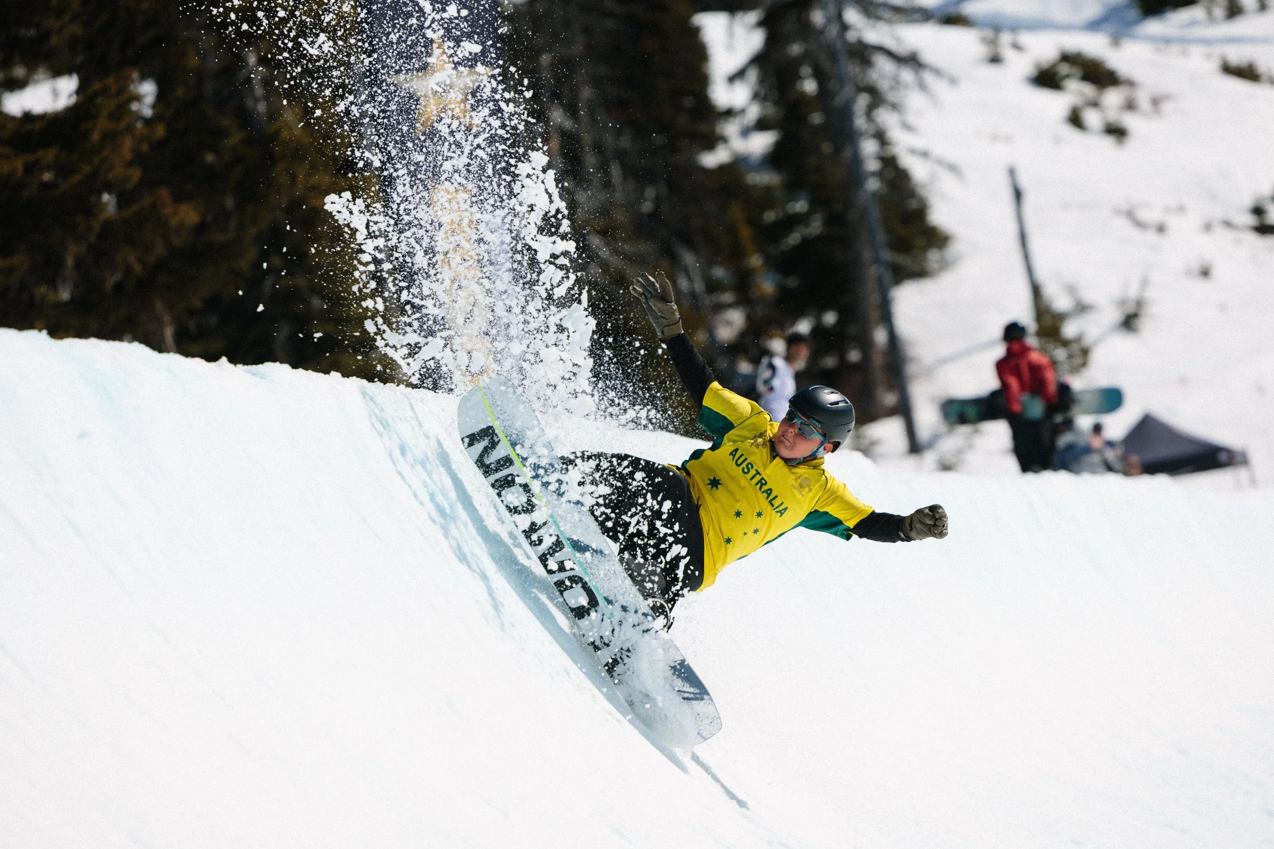 A snowboarder wearing a yellow jacket with 'Australia' written on it, black helmet, and sunglasses, is falling in the snow on a snowy slope with trees in the background.