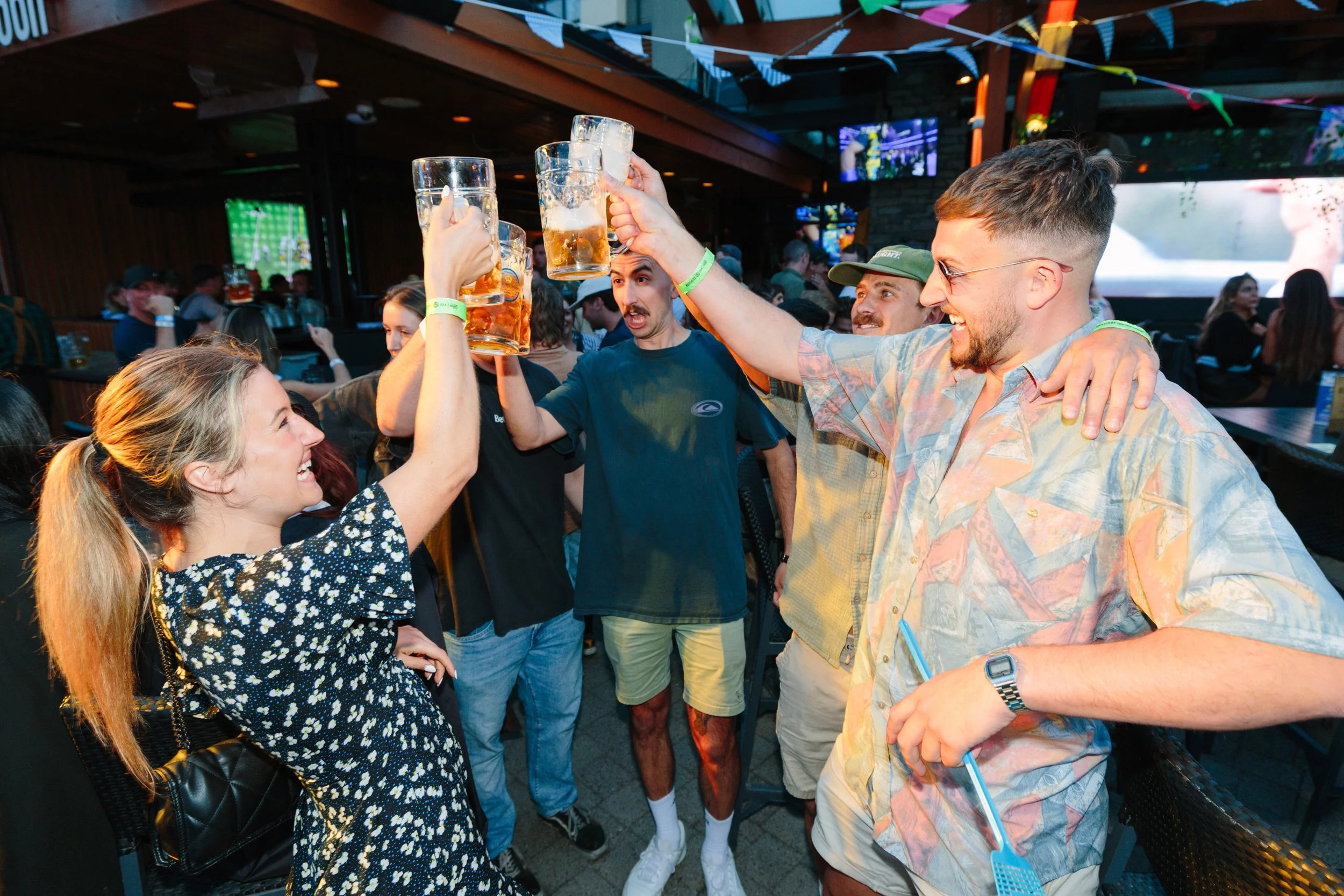 A group of friends celebrating with beer mugs at a lively bar or pub with colorful banners overhead and a bustling crowd in the background.
