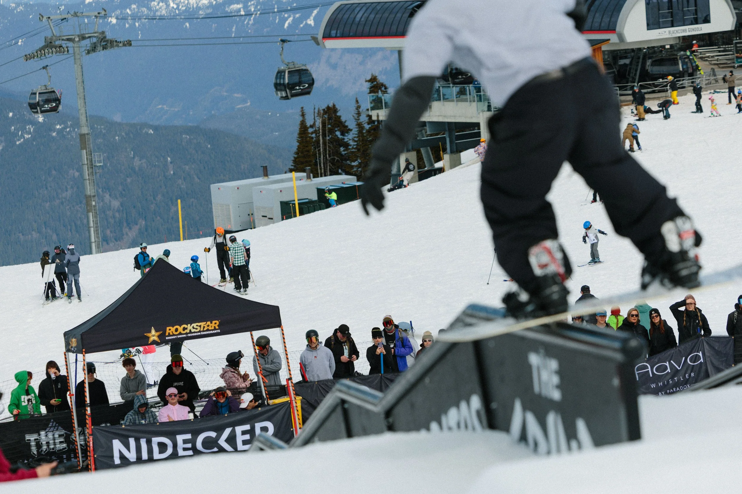 People skiing and snowboarding on a snowy mountain at a ski resort. In the background, there are ski lifts, a ski lodge, and a crowd of visitors. There is a black tent with yellow logo, and people in colorful winter gear.