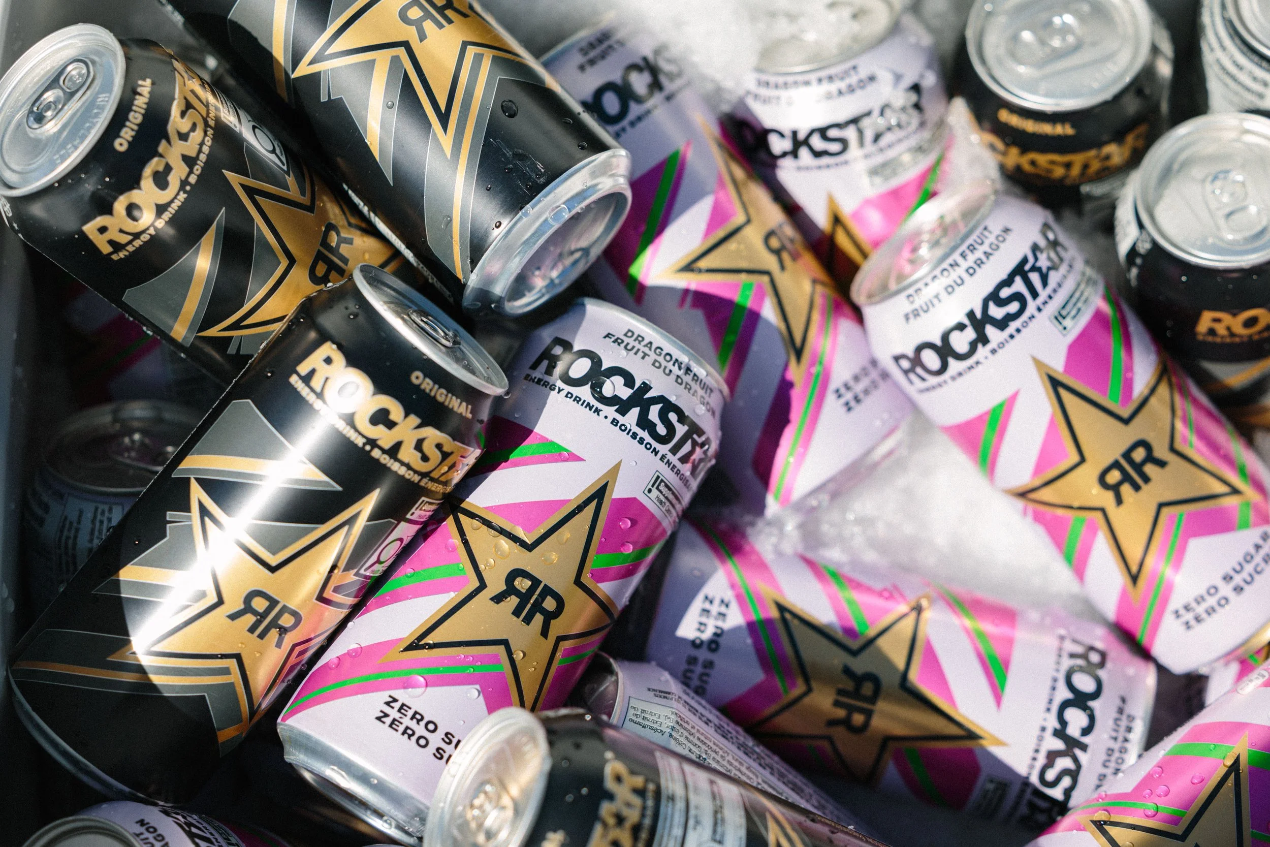 A pile of Rockstar energy drink cans, some in black and gold packaging and others in pink and white packaging, with condensation droplets on the cans.