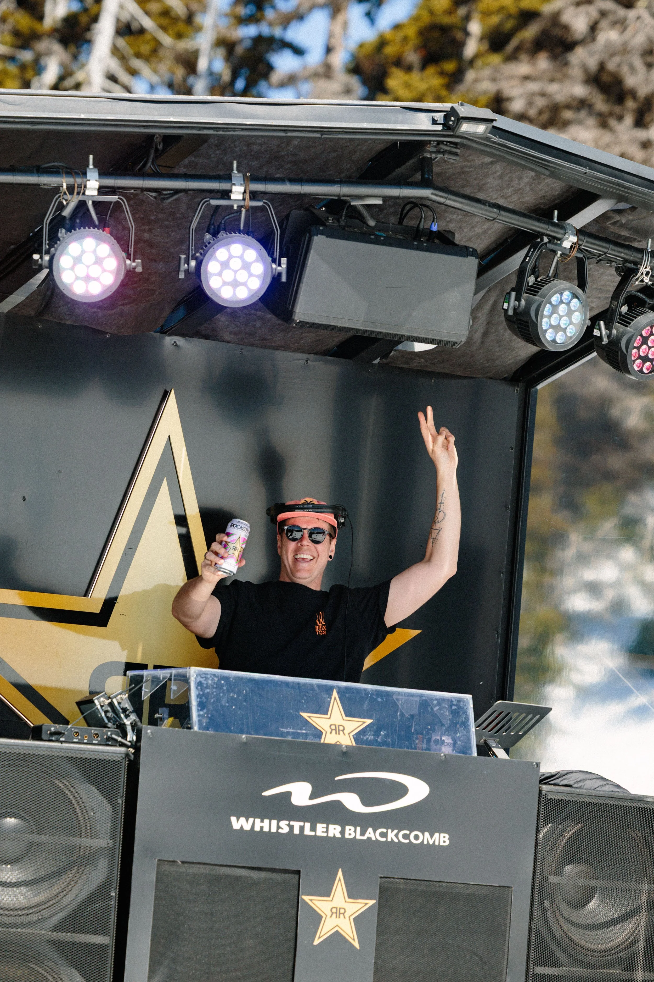 A DJ is standing behind a DJ booth at an outdoor event, wearing sunglasses and a black t-shirt, holding a can of drink, and making a peace sign with his right hand. The booth has the logo of Whistler Blackcomb and a gold star with the initials 'RR' o