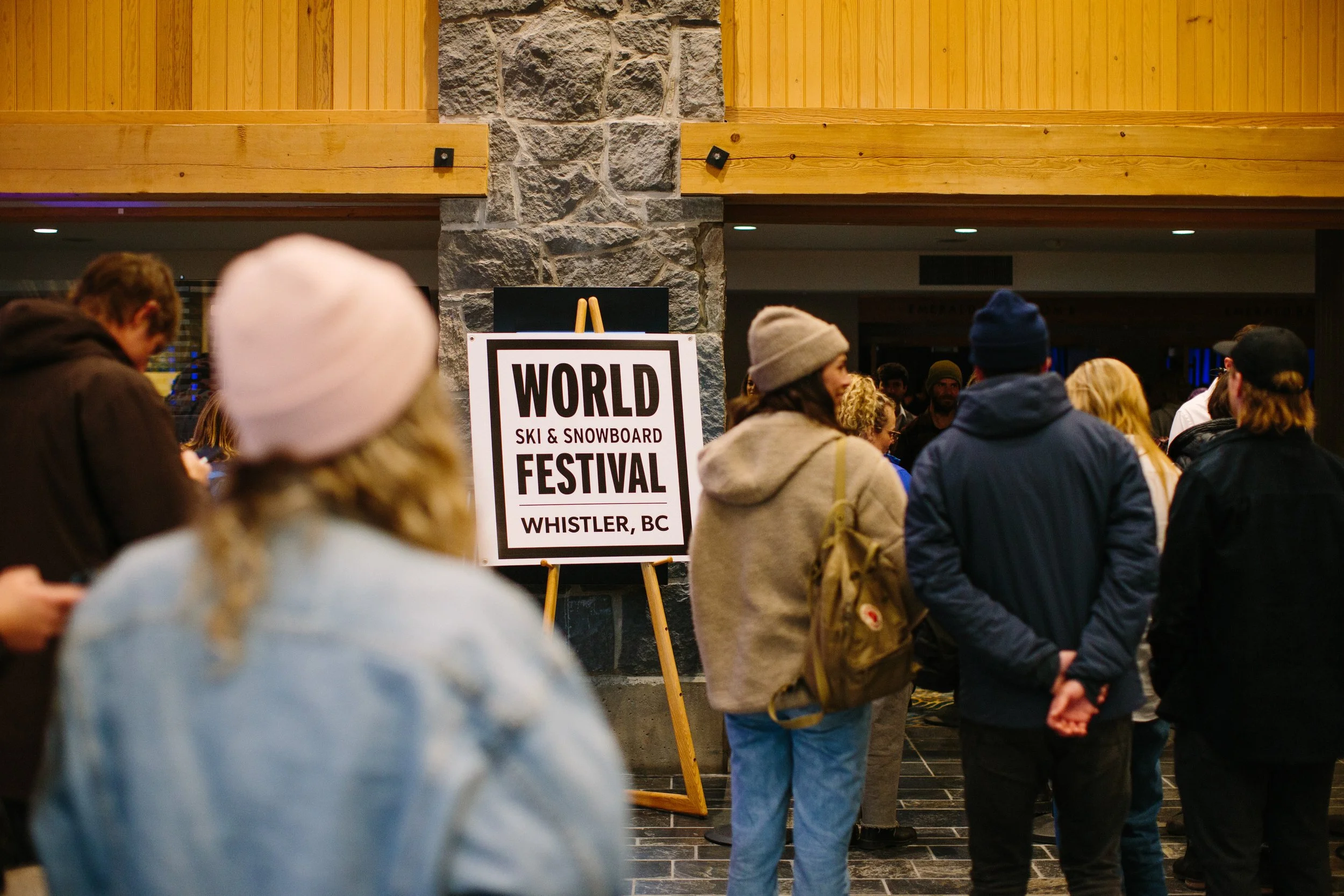A crowd of people gathered indoors at the World Ski & Snowboard Festival in Whistler, BC, with some wearing winter clothing and hats, near a sign and a stone and wood interior wall.