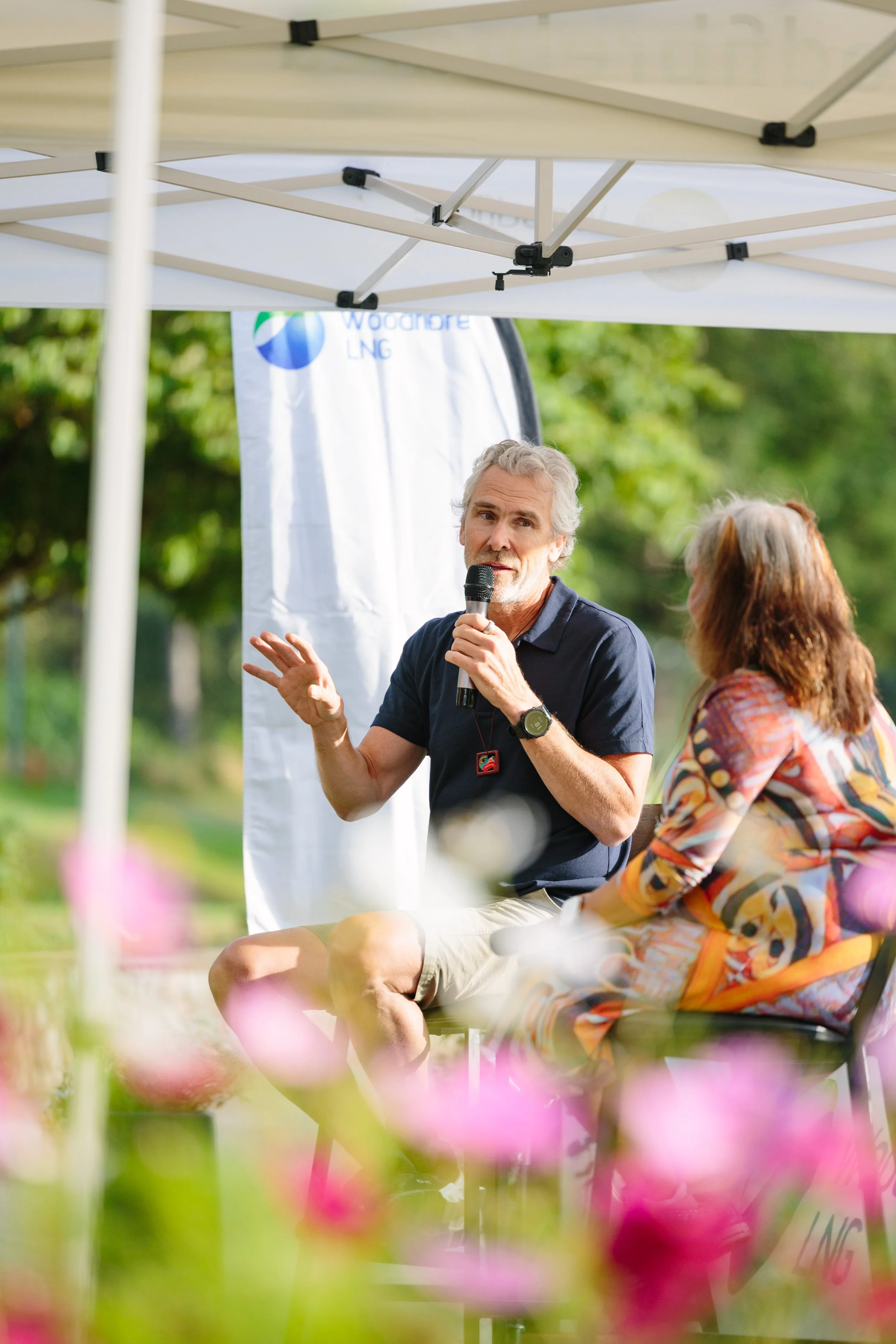 A man in a dark polo shirt and shorts sitting under a white outdoor tent, speaking into a microphone during an outdoor event, with a woman seated beside him and green trees in the background.