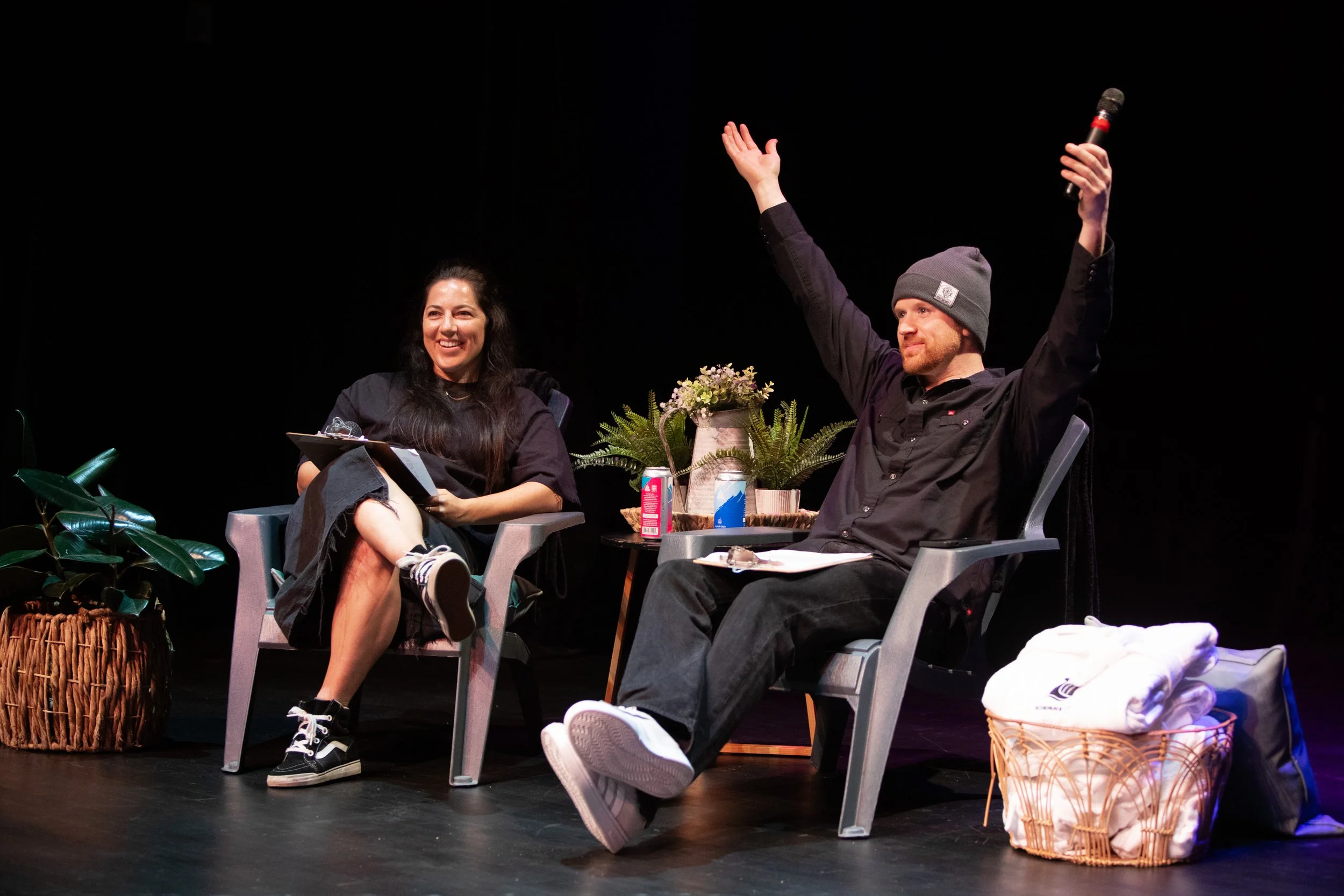 A woman and a man sitting in chairs on stage, smiling and engaging in conversation. The woman has long dark hair, and the man is wearing a gray beanie and holding a microphone. There are plants, a small table with drinks, and a basket of towels nearb