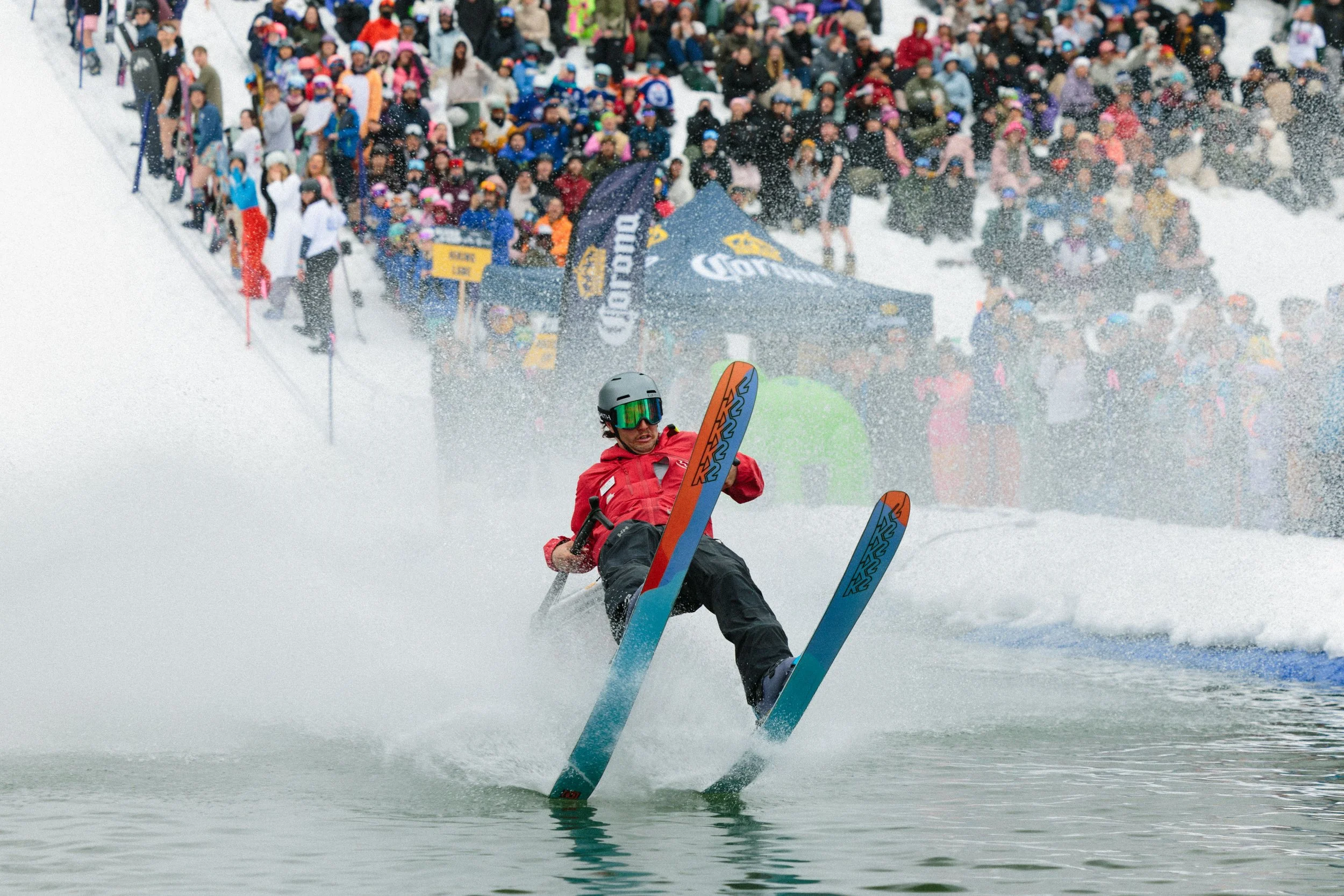A person in a red jacket and dark pants is skiing on water, wearing a helmet and goggles, with a large crowd of skiers and spectators gathered on snow in the background.