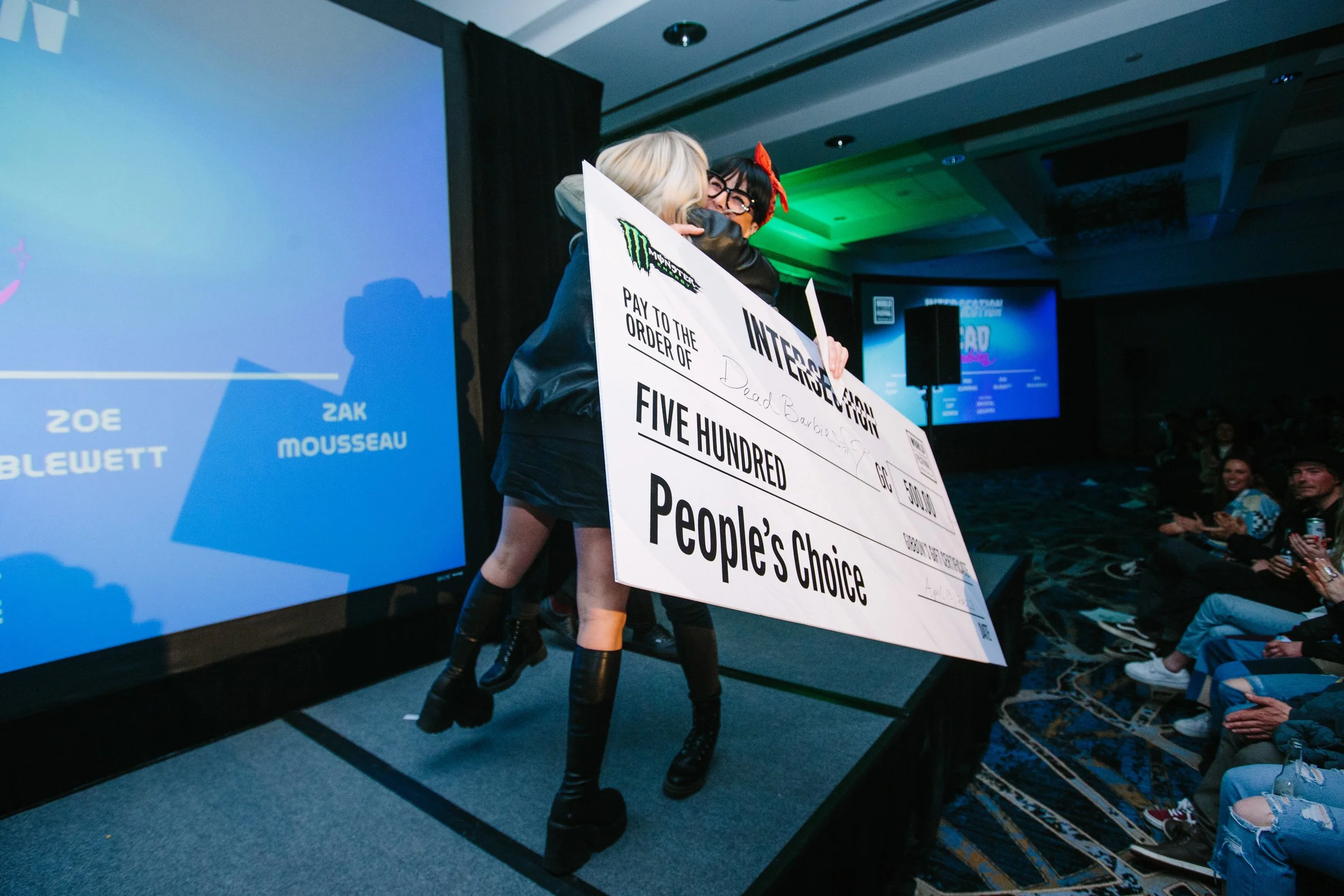 Two individuals in cosplay costumes hugging on stage during an event, holding an oversized check for five hundred people’s choice award, with audience members watching and clapping in the background.