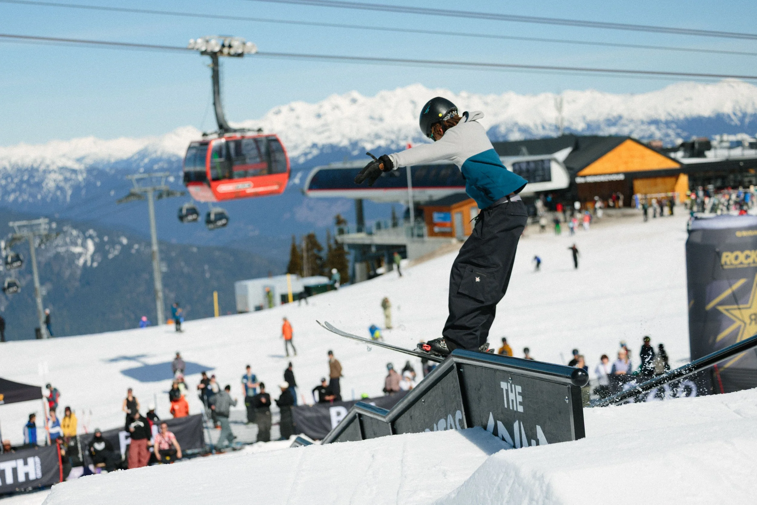 A person snowboarding down a ramp at a ski resort with a snowy mountain landscape in the background and a crowd of spectators.