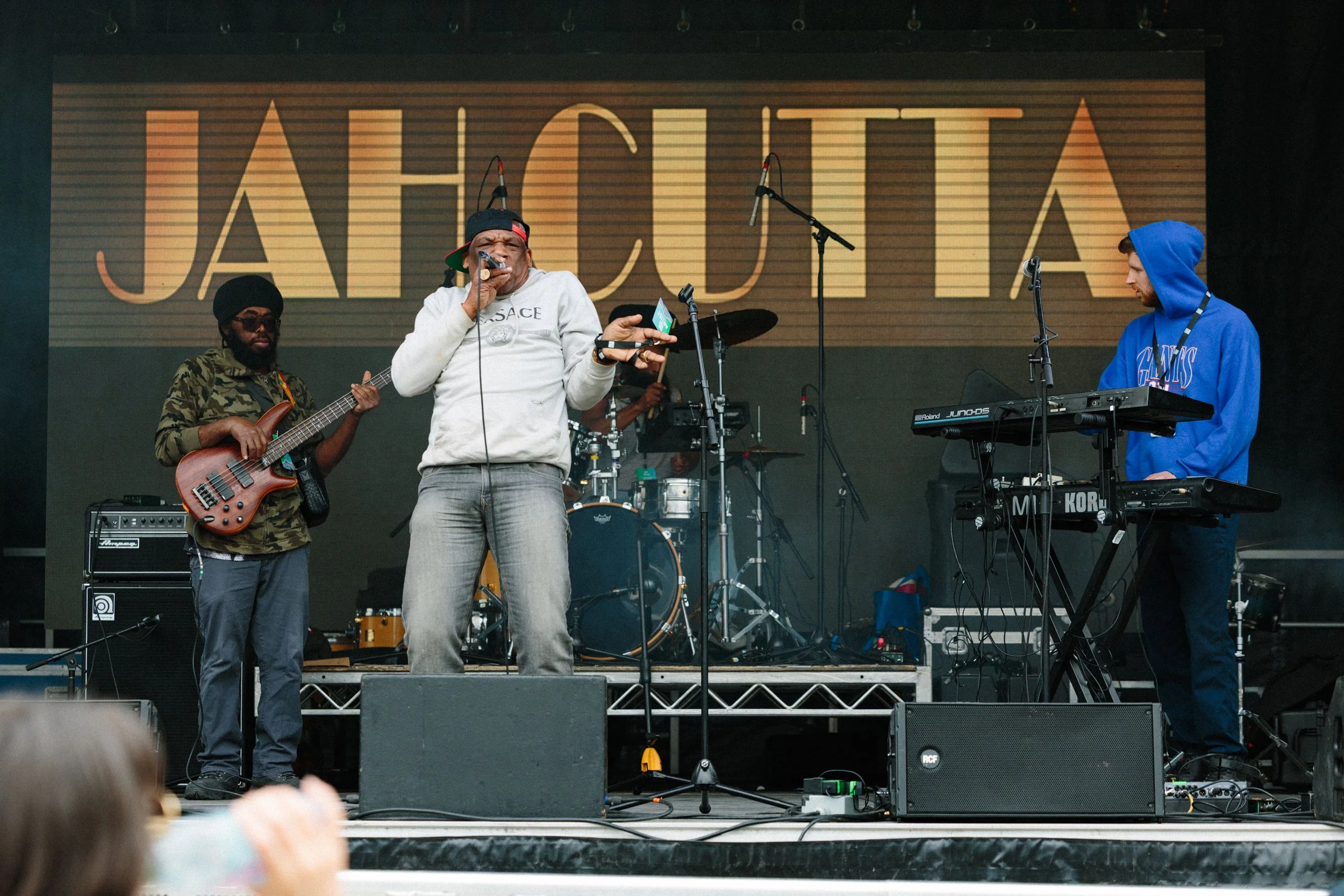 A music band is performing on stage with a large screen in the background displaying the words "JAH CULTA". The band includes a person singing with a microphone, a guitarist, a keyboardist, and a drummer. There are music equipment and microphones on 