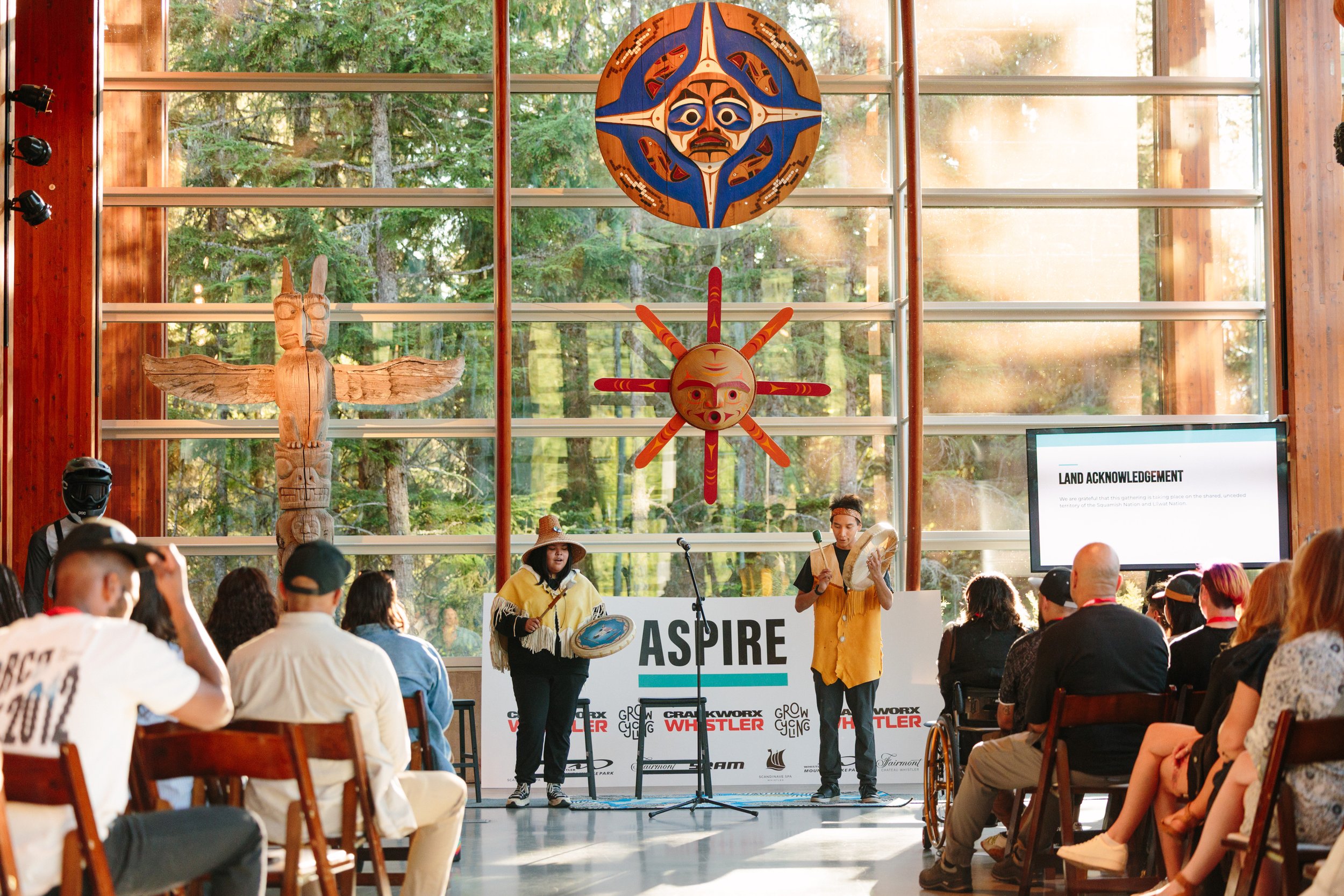 Indoor cultural event featuring performers in traditional attire with musical instruments, seated audience watching, large wooden totem pole, Native art masks and decorations on the wall, and a digital screen displaying a land acknowledgement.