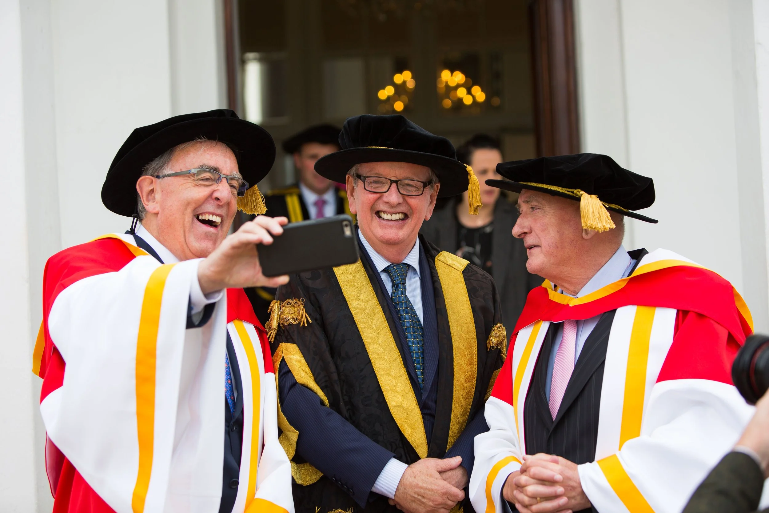 Three academics in graduation regalia taking a selfie at a graduation ceremony, with people and decorations in the background.