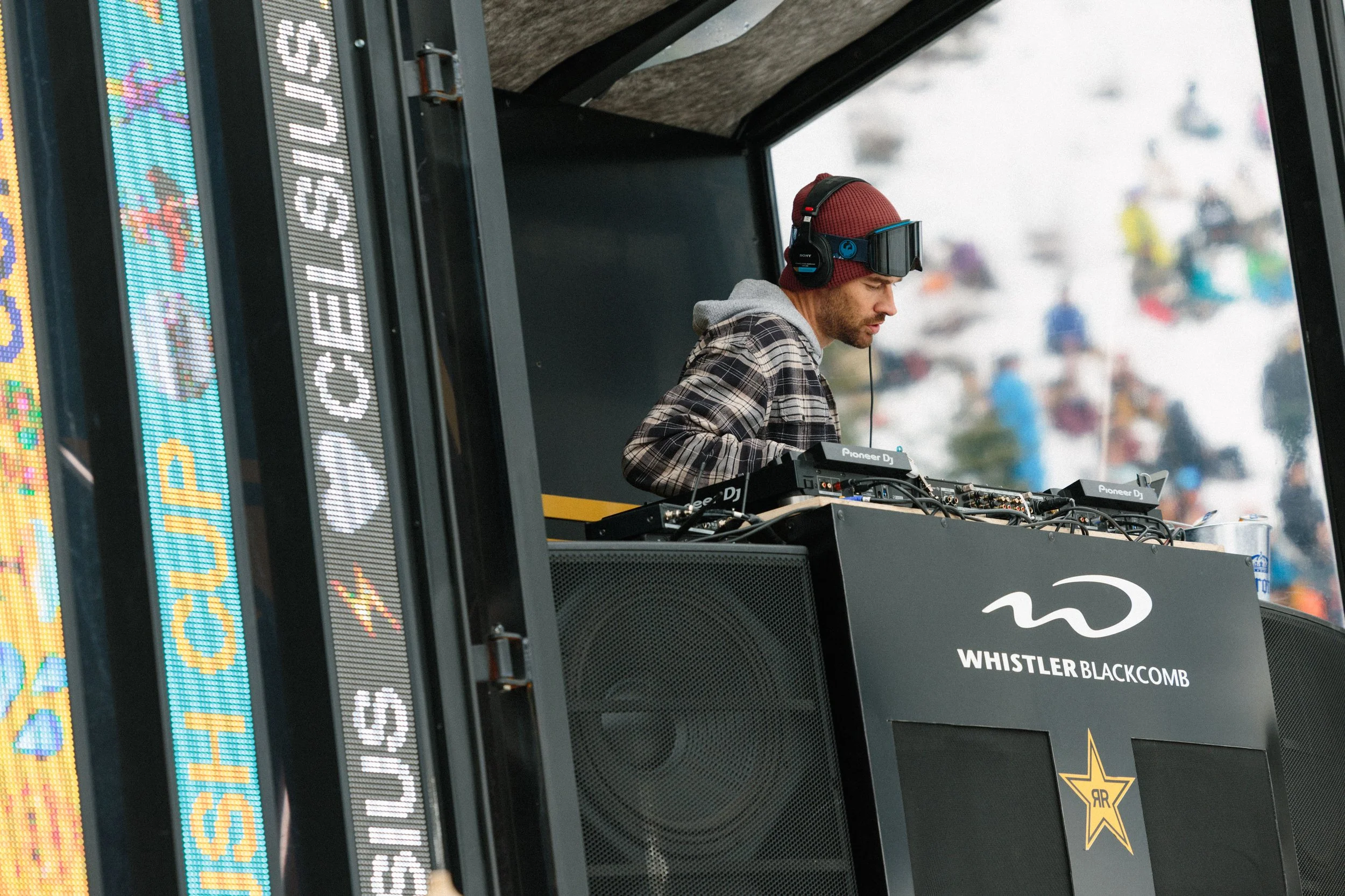 A male DJ wearing a red beanie, gray hoodie, and checkered shirt, with headphones and goggles, performing at an outdoor music event with digital screens in the background displaying colorful graphics and the words "Whistler Blackcomb."