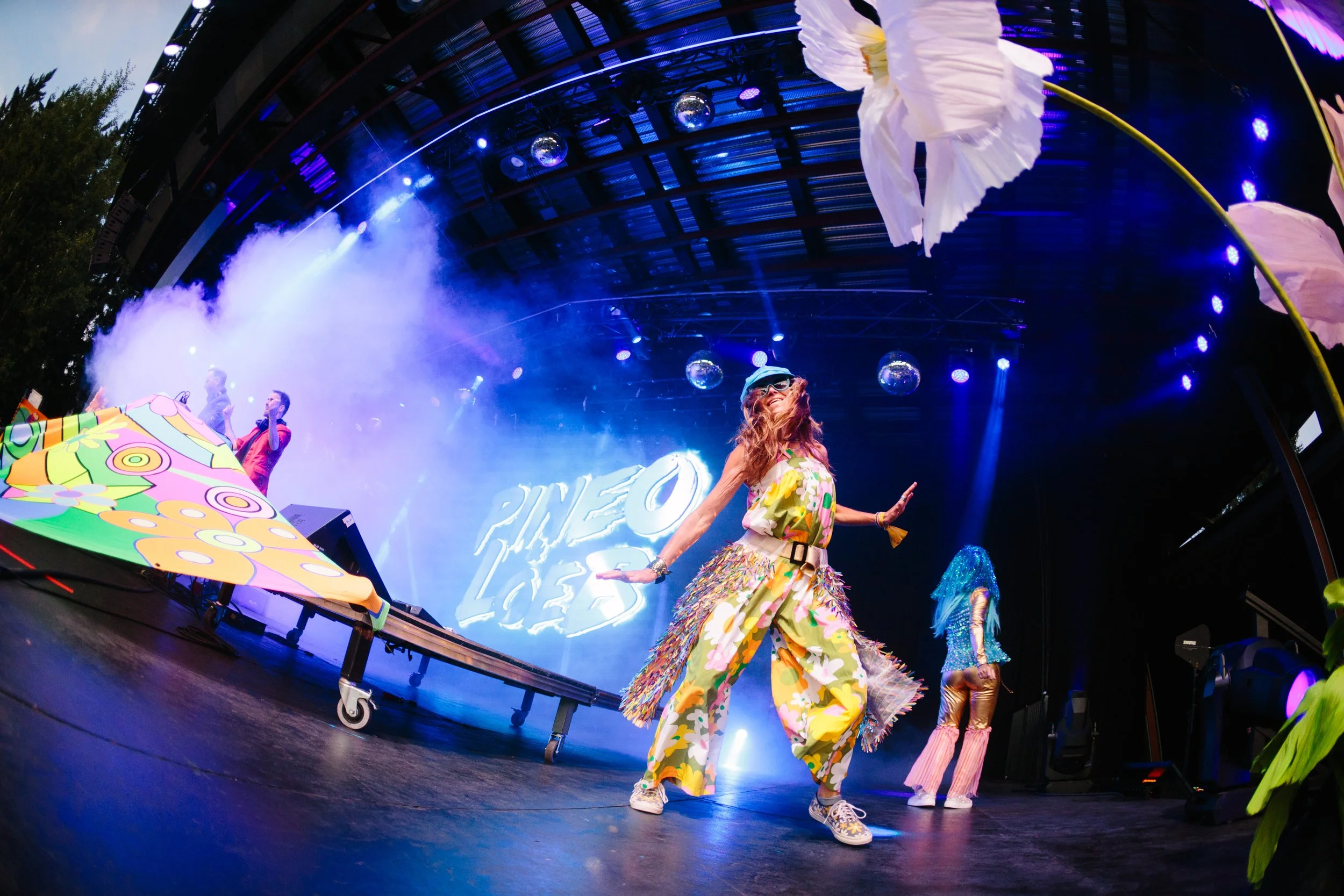 Colorful performers dancing on stage with vibrant costumes and psychedelic backdrop at PINEO 2023 event with colorful props and stage lights.