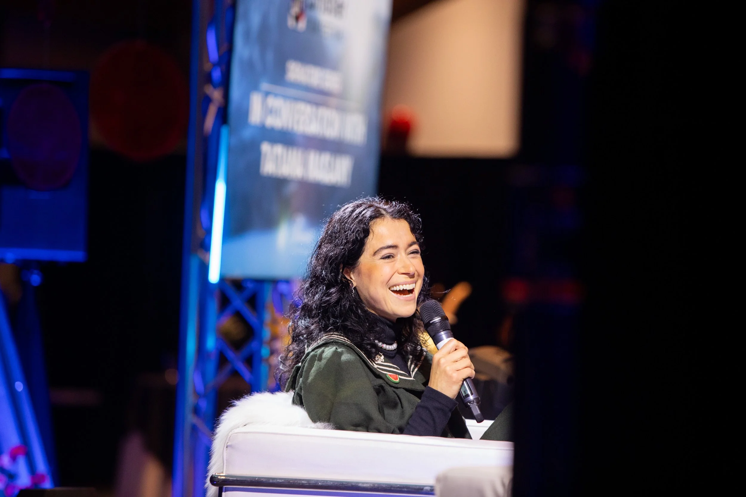 A woman with curly black hair smiling and speaking into a microphone at an event, sitting on a white chair with a backdrop screen and stage equipment behind her.