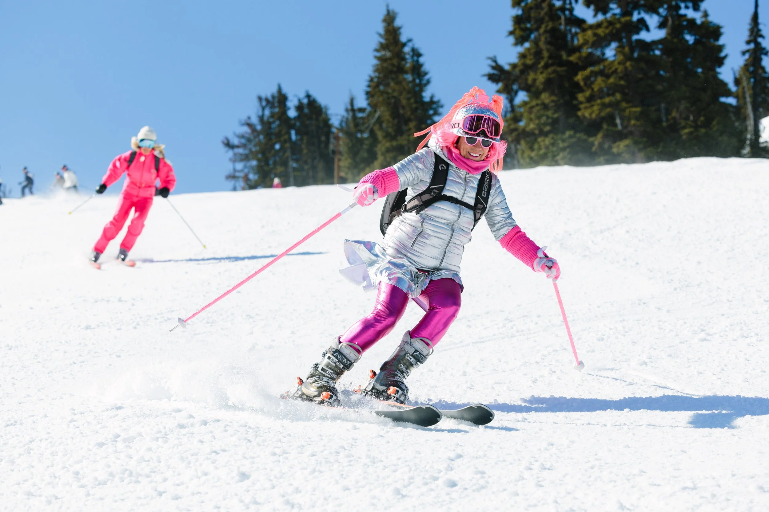 Two children skiing on a snowy slope, wearing pink and silver winter outfits, with a backdrop of green pine trees and a bright blue sky.