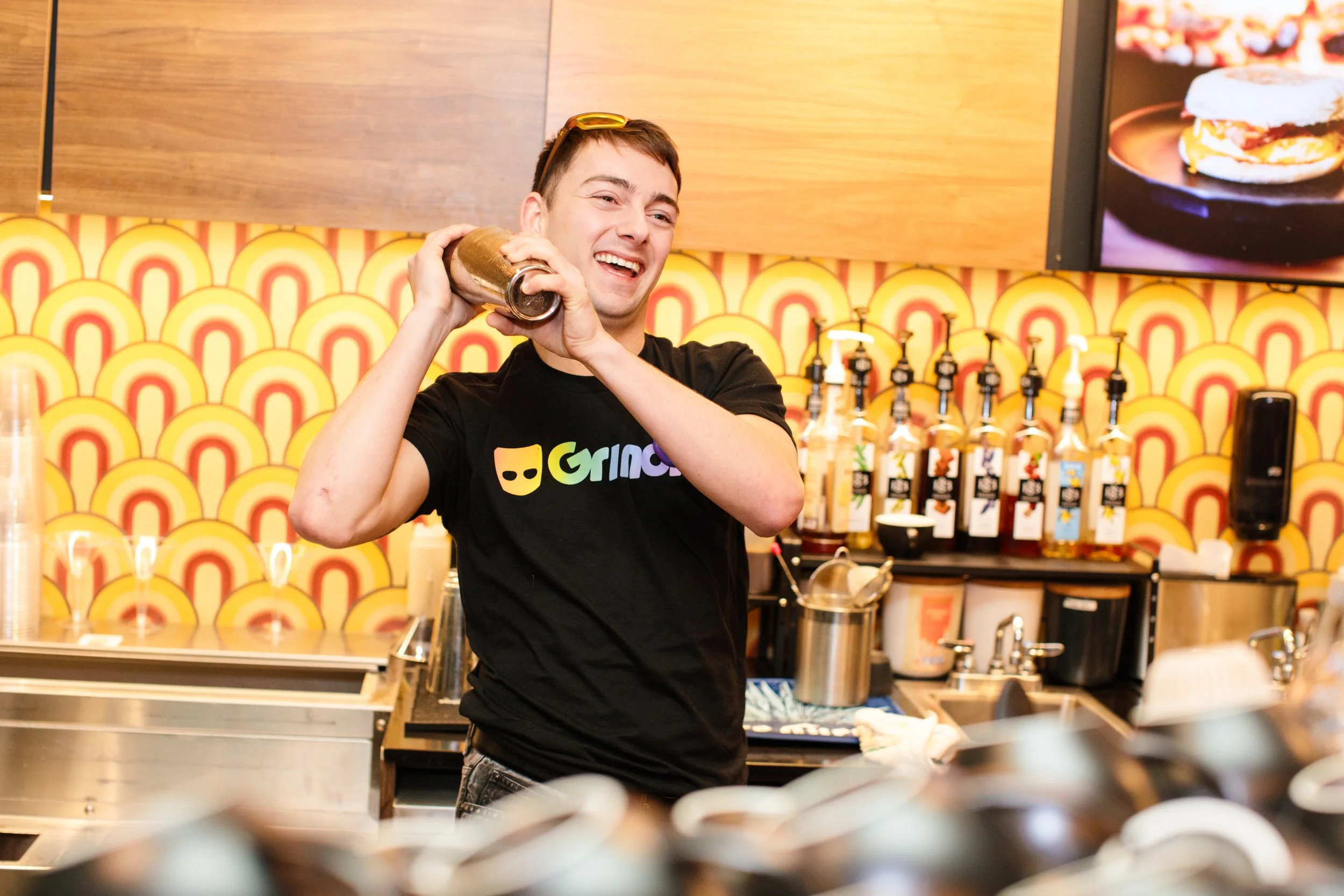 A smiling bartender in a black shirt with a colorful logo, holding a cocktail shaker to his shoulder inside a lively bar with yellow and orange patterned wallpaper and a TV screen showing a sandwich.