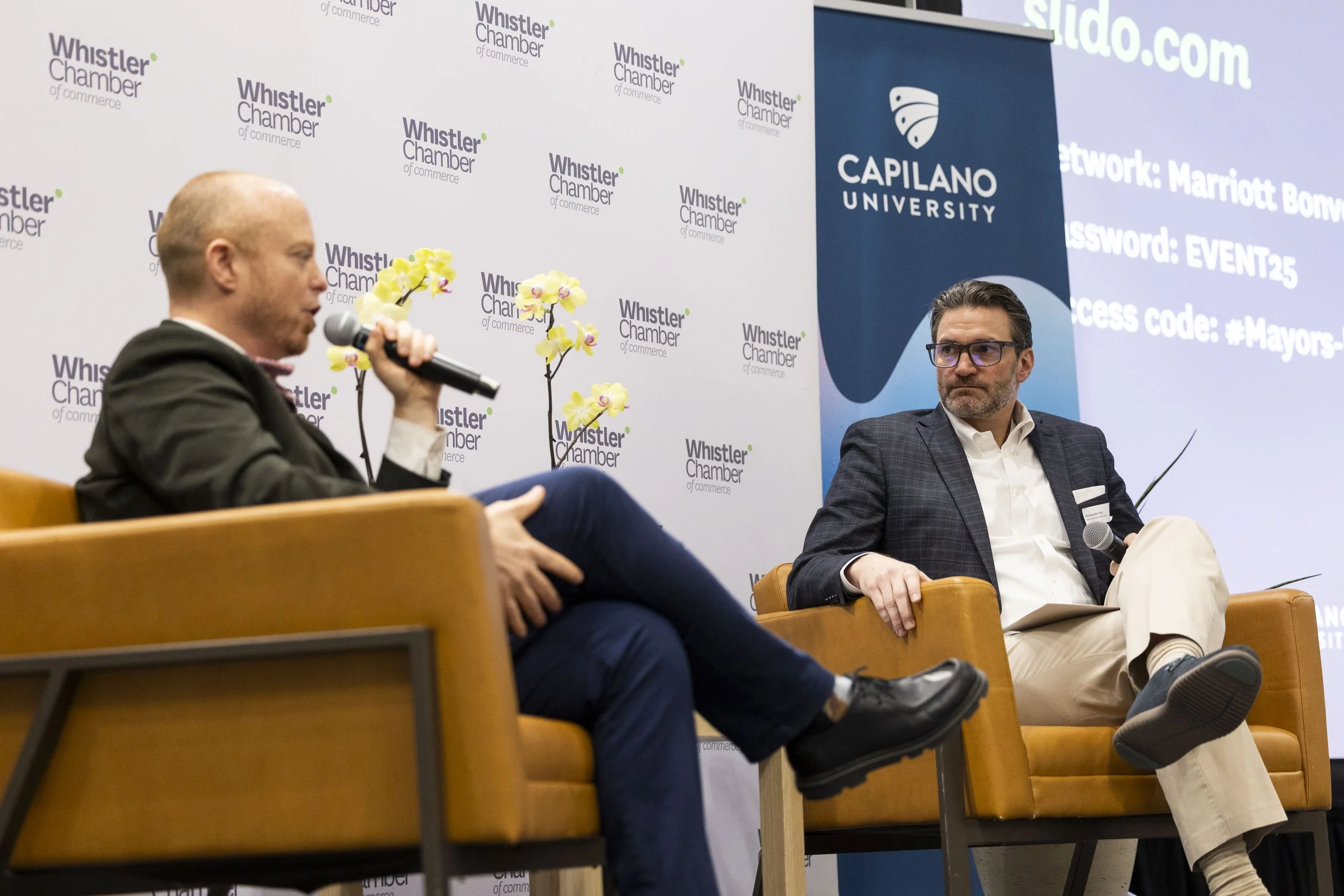 Two men engaged in a conversation on stage during a panel discussion, seated in orange chairs with a Whistler Chamber of Commerce backdrop and a blue Capilano University banner behind them. One man is speaking into a microphone, and the other man is 