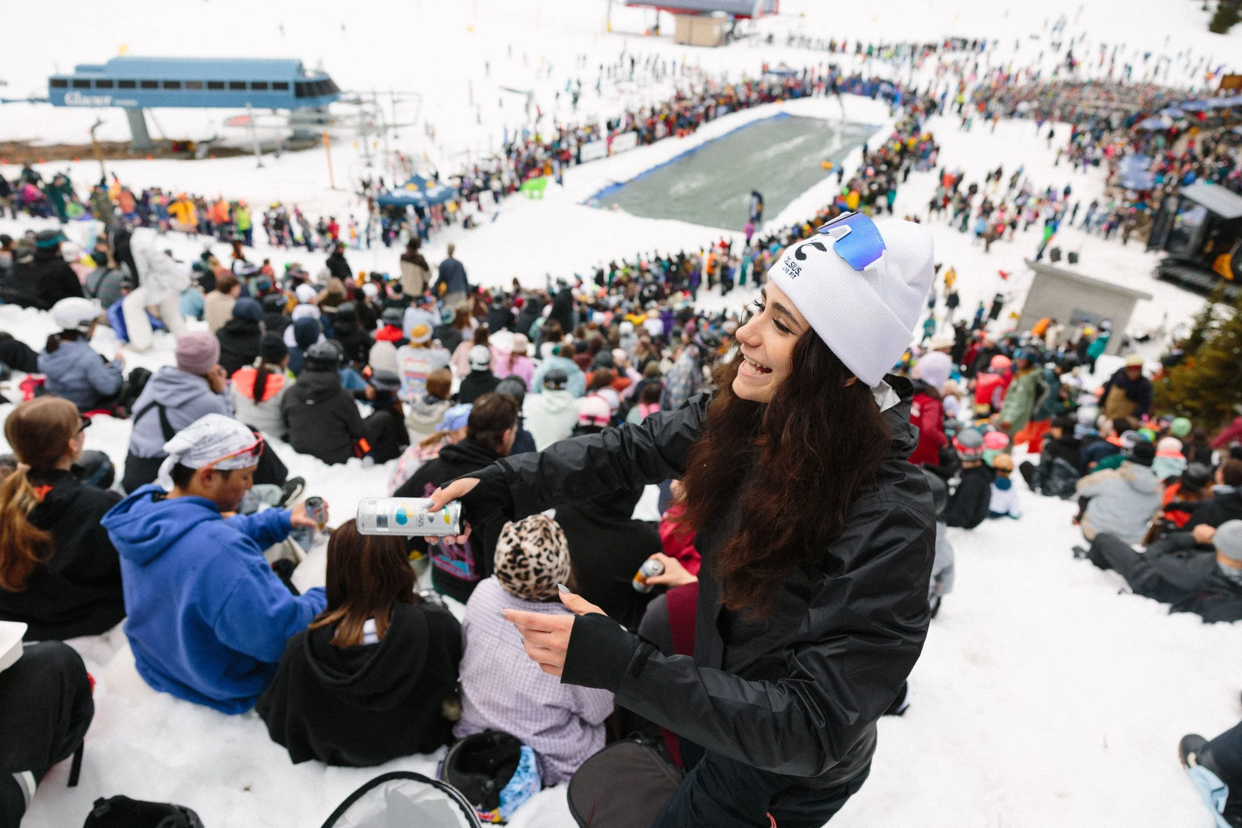 Crowd gathered on snowy ground at a ski resort, with many people sitting and standing, some holding drinks, and a smiling young woman in the foreground wearing a white beanie and goggles.