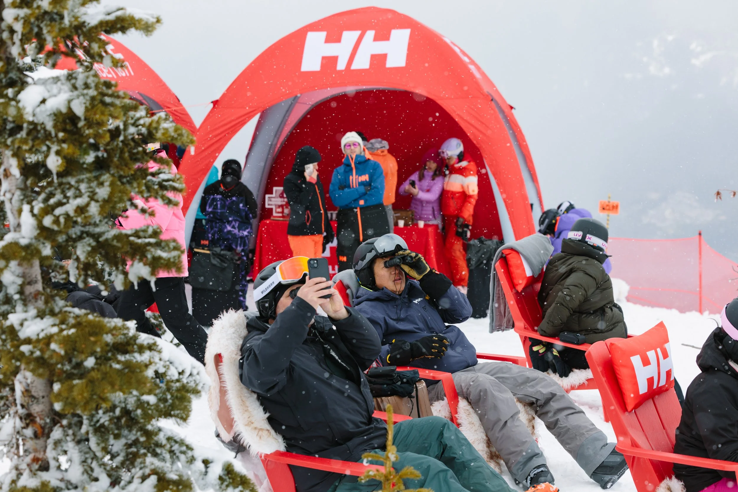 People sitting in red sleds on snow near a red tent with snow-covered trees in the background. The scene appears to be at a ski resort.