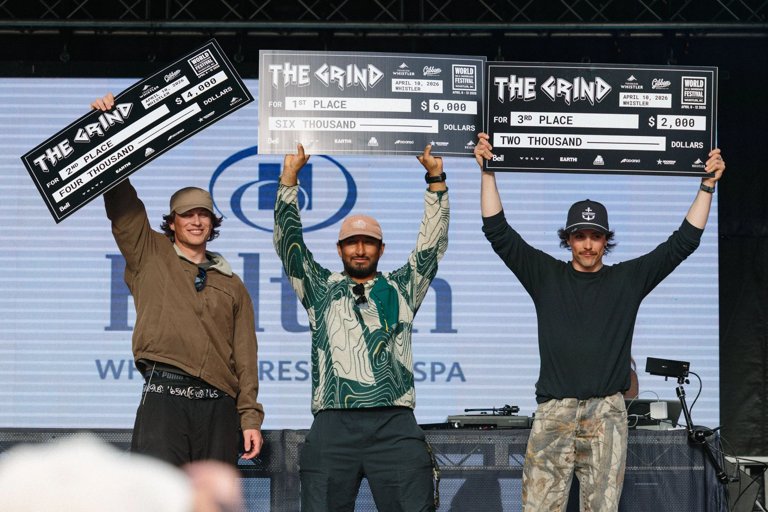 Three men standing on a stage holding oversized checks for winning prizes at an event called The Grind. The checks are for 1st place, 2nd place, and 3rd place, with monetary amounts of $6,000, $4,000, and $2,000 respectively. There is a large screen 