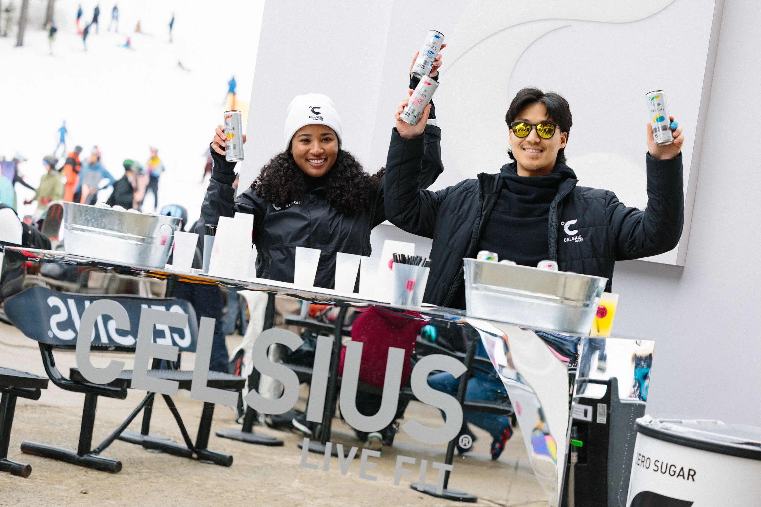 Two people smiling and holding cans of Celsius energy drink at an outdoor event, with a table of promotional materials and a crowd of people in the snowy background.