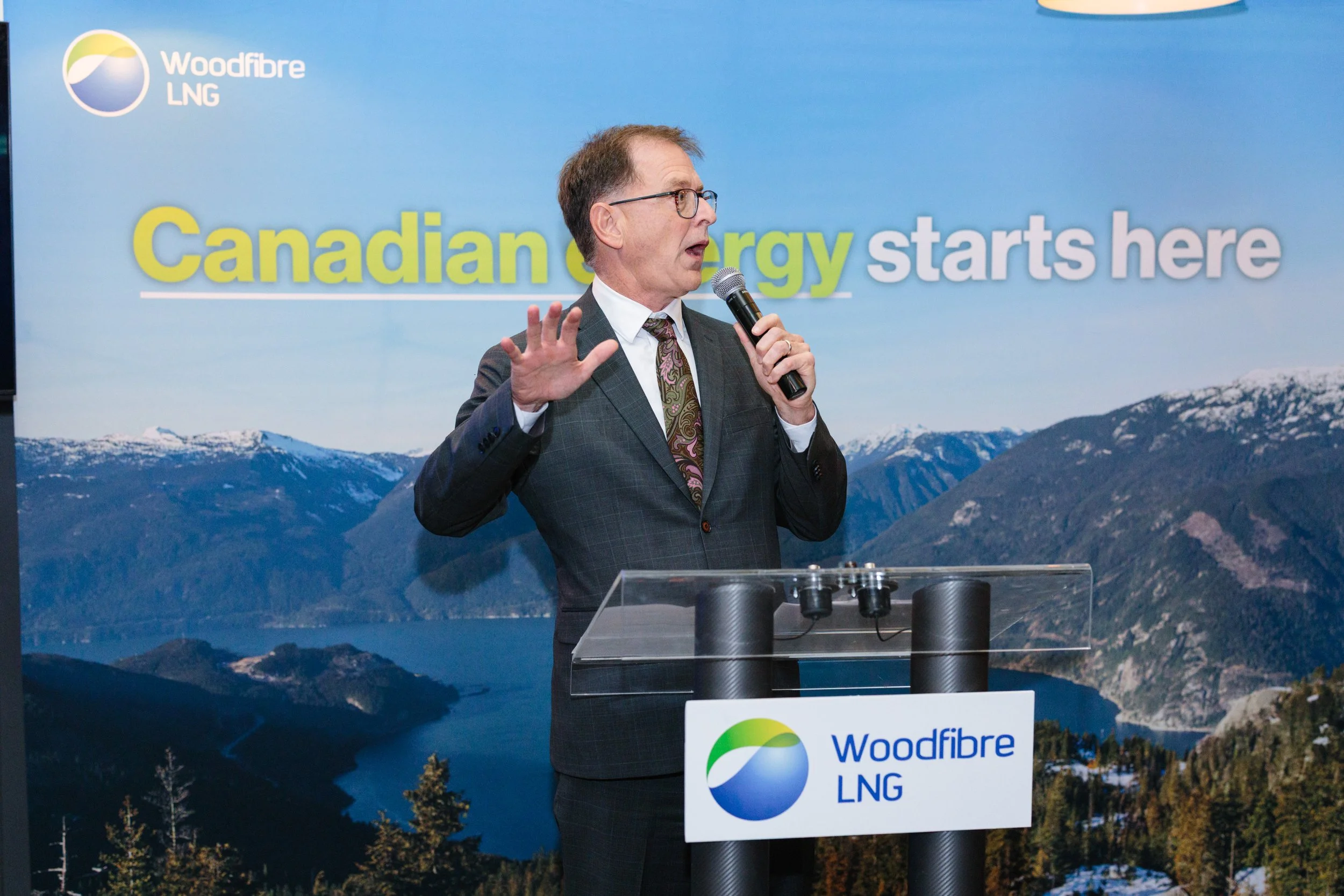 A man in a suit with glasses holding a microphone and gesturing with his hand while speaking at a podium with a "Woodfibre LNG" sign, in front of a scenic backdrop of mountains and a lake, with the text "Canadian energy starts here" displayed in the 