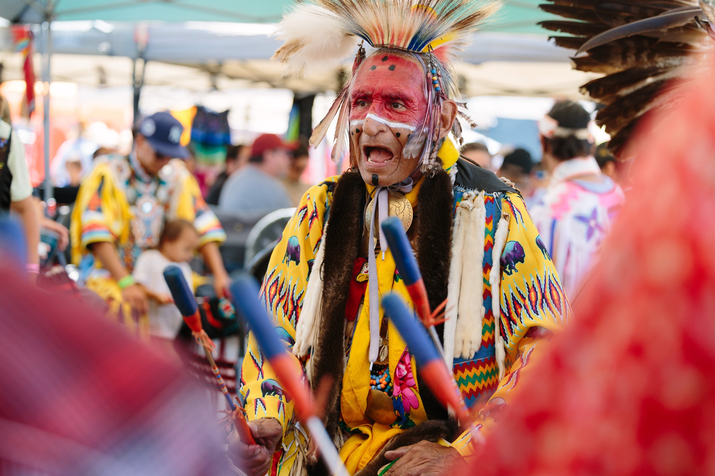 Man dressed in traditional Native American clothing with face paint, feathers, and a yellow robe, participating in a cultural event or celebration.