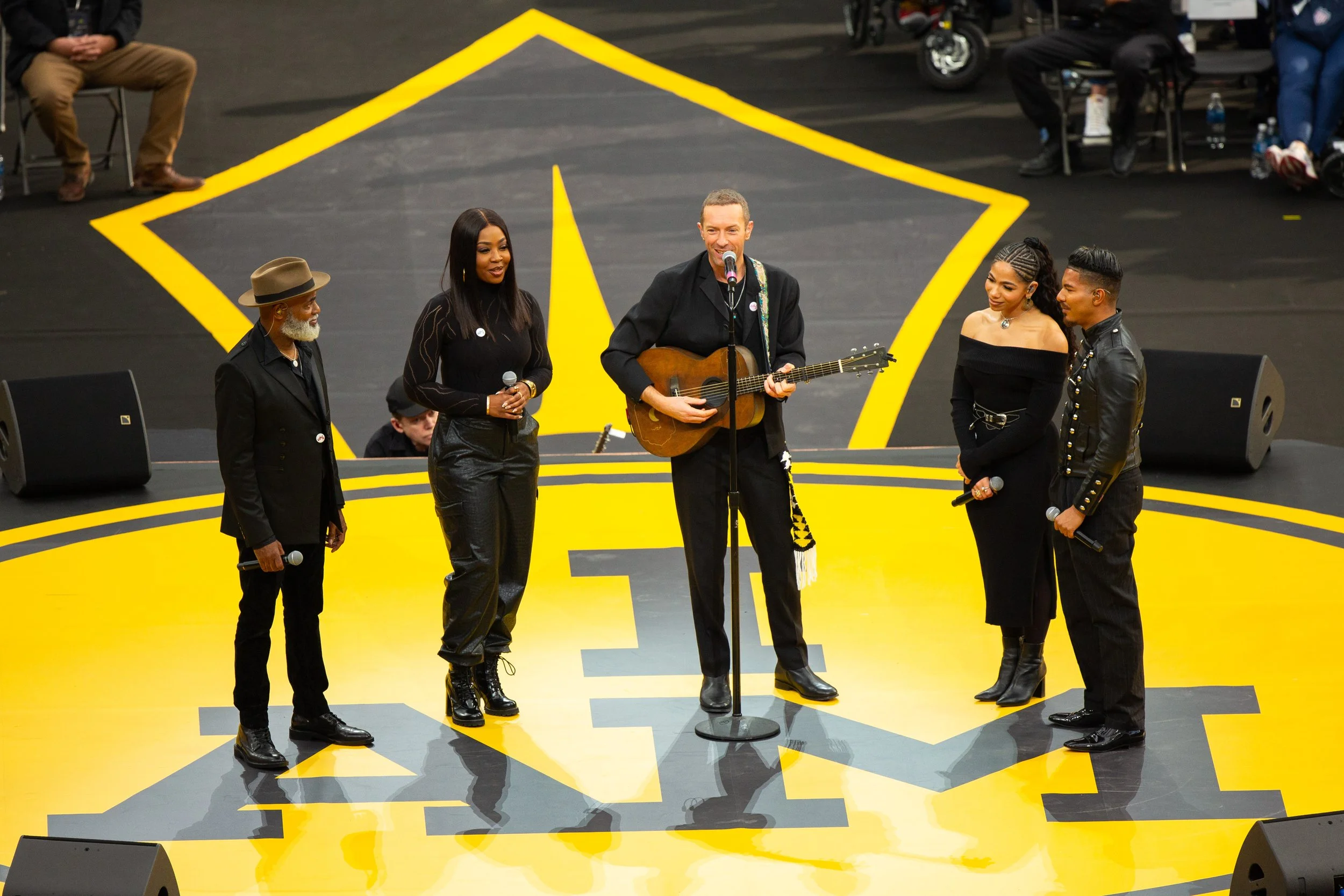 A musical performance on a yellow and black stage with five singers, one playing guitar, surrounded by audience members and speakers.