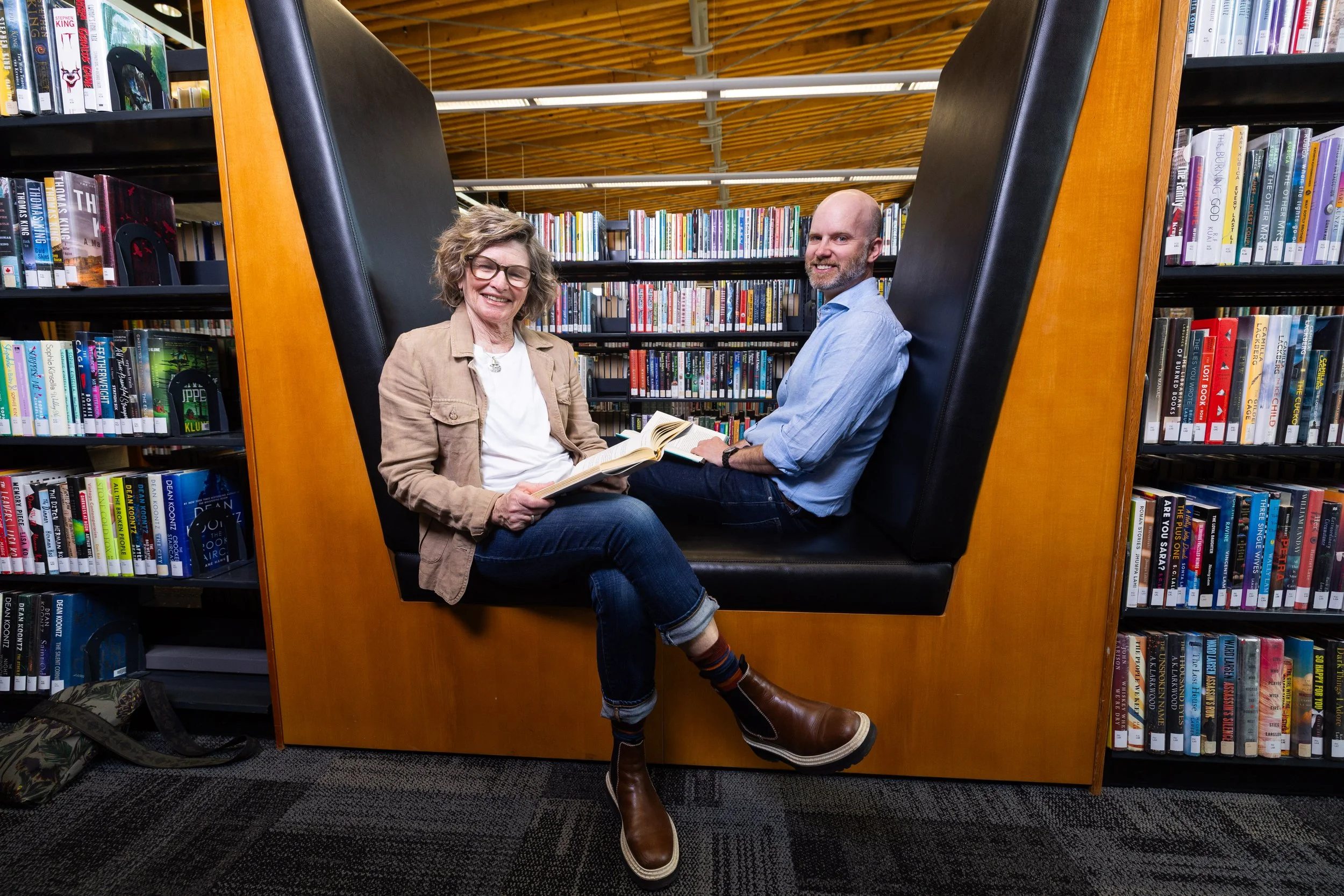 Two people sitting inside a large black booth in a library, smiling and reading books, with shelves of books behind them.