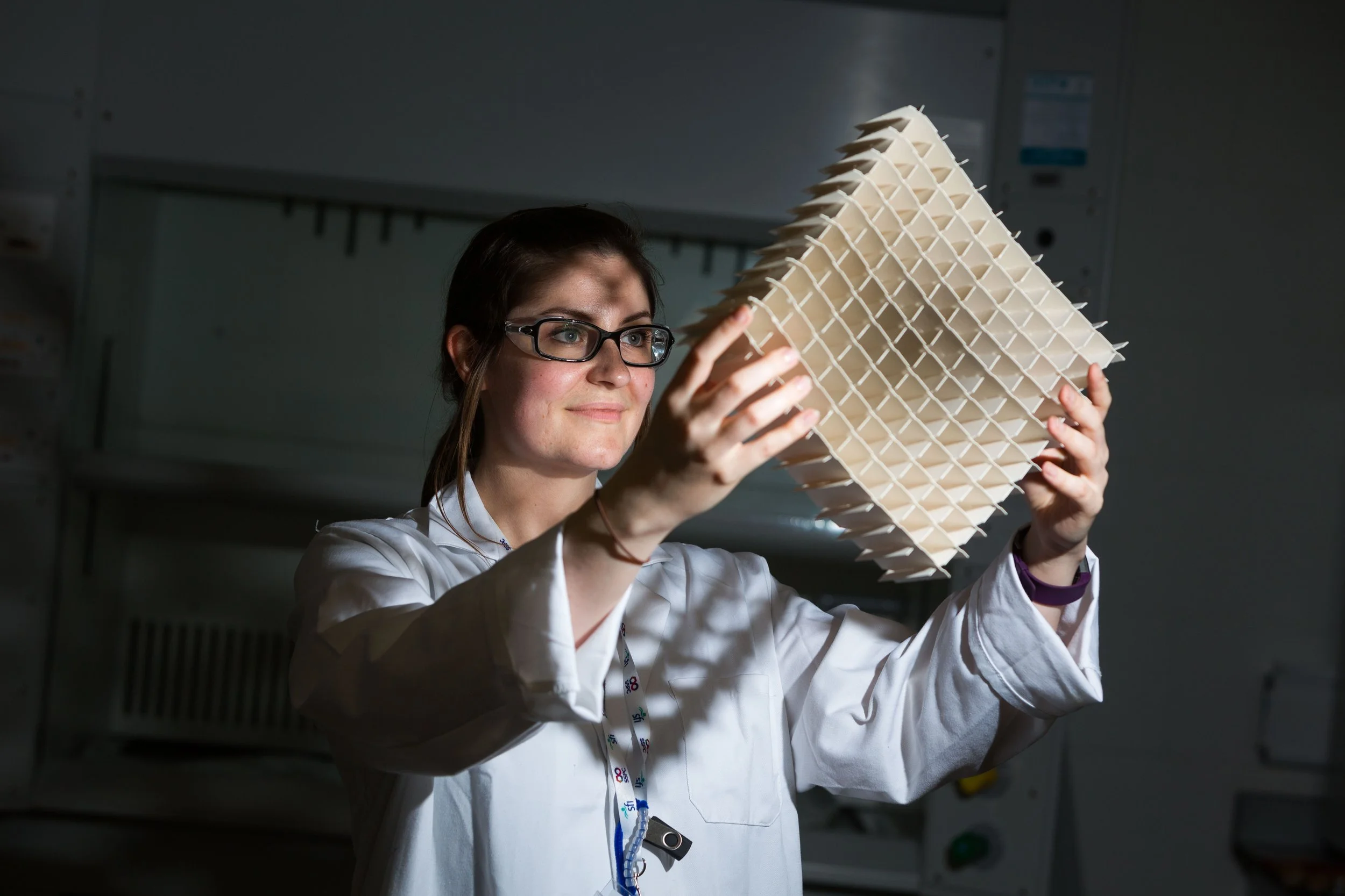 A scientist wearing glasses and a white lab coat holds up a large, three-dimensional geometric structure made of interconnected paper or plastic grid panels in a laboratory setting.