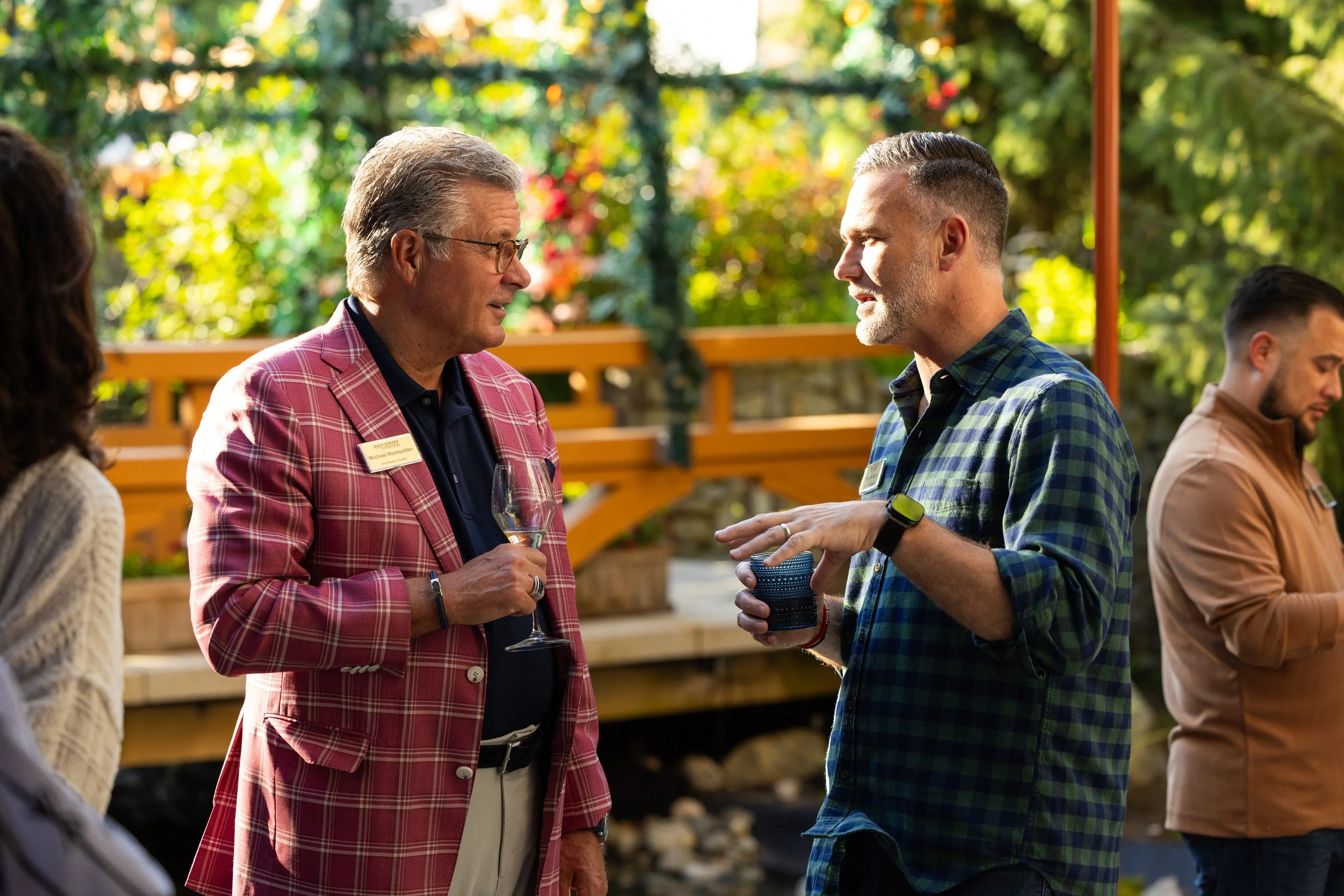 Two men are conversing at an outdoor gathering on a sunny day. One man is wearing glasses, a red plaid blazer, and holding a glass of wine. The other man is wearing a blue and green plaid shirt, holding a cup, and wearing a watch. There are trees and