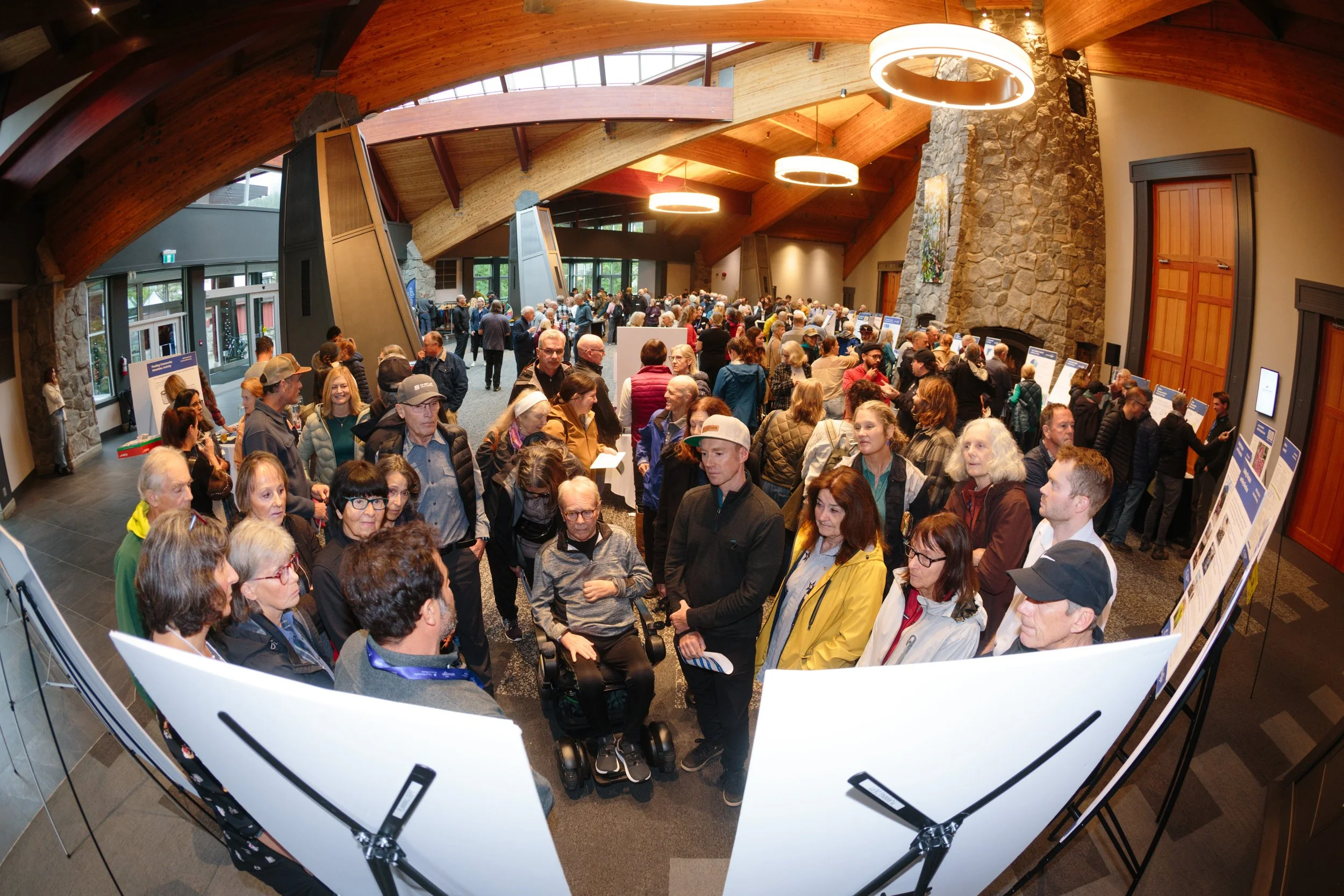 Crowd of people attending an indoor event, many gathered around display boards, in a large room with high wooden vaulted ceilings and stone walls.