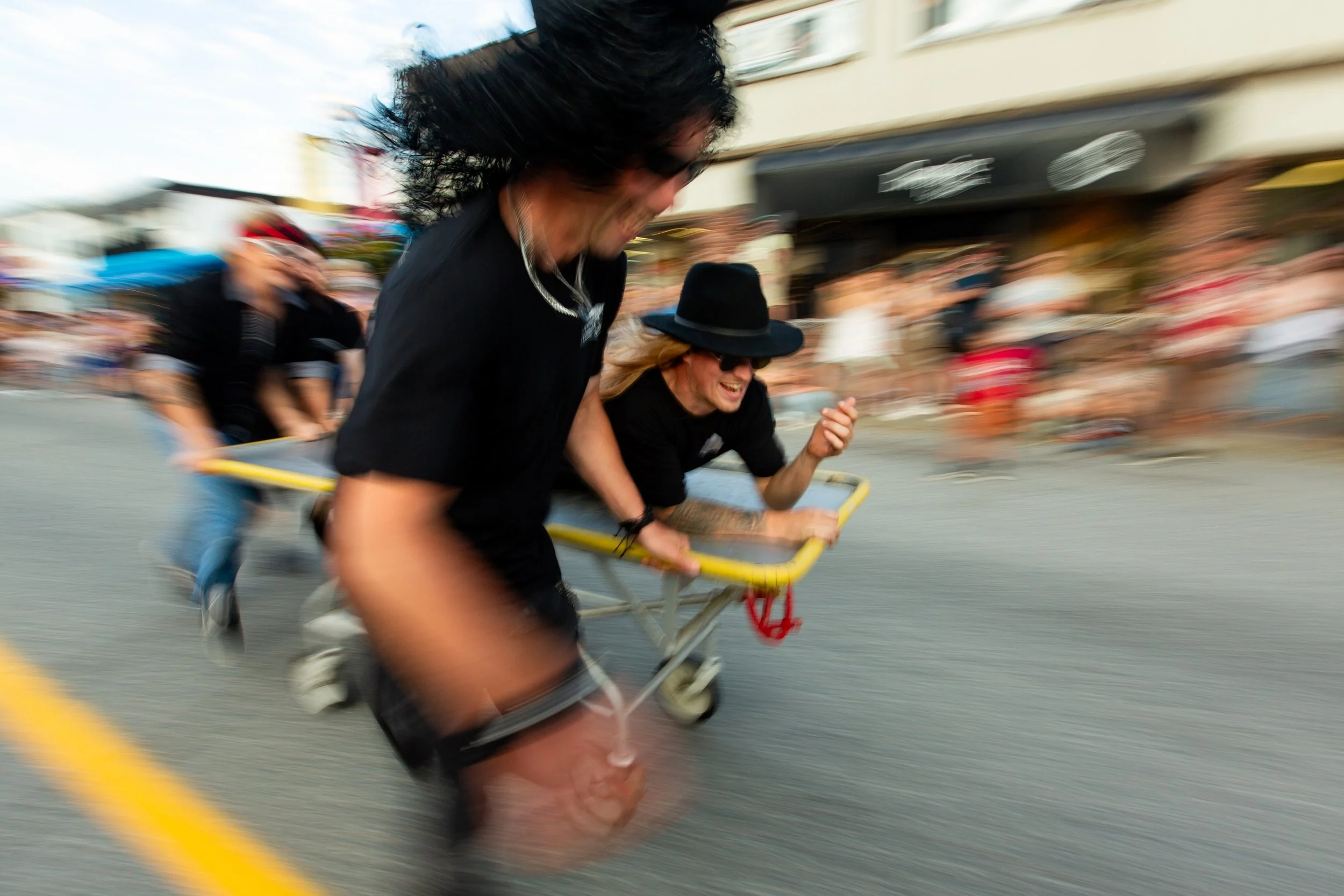 Two people crashing on a yellow street cart during a race, with a crowd of spectators blurred in the background.