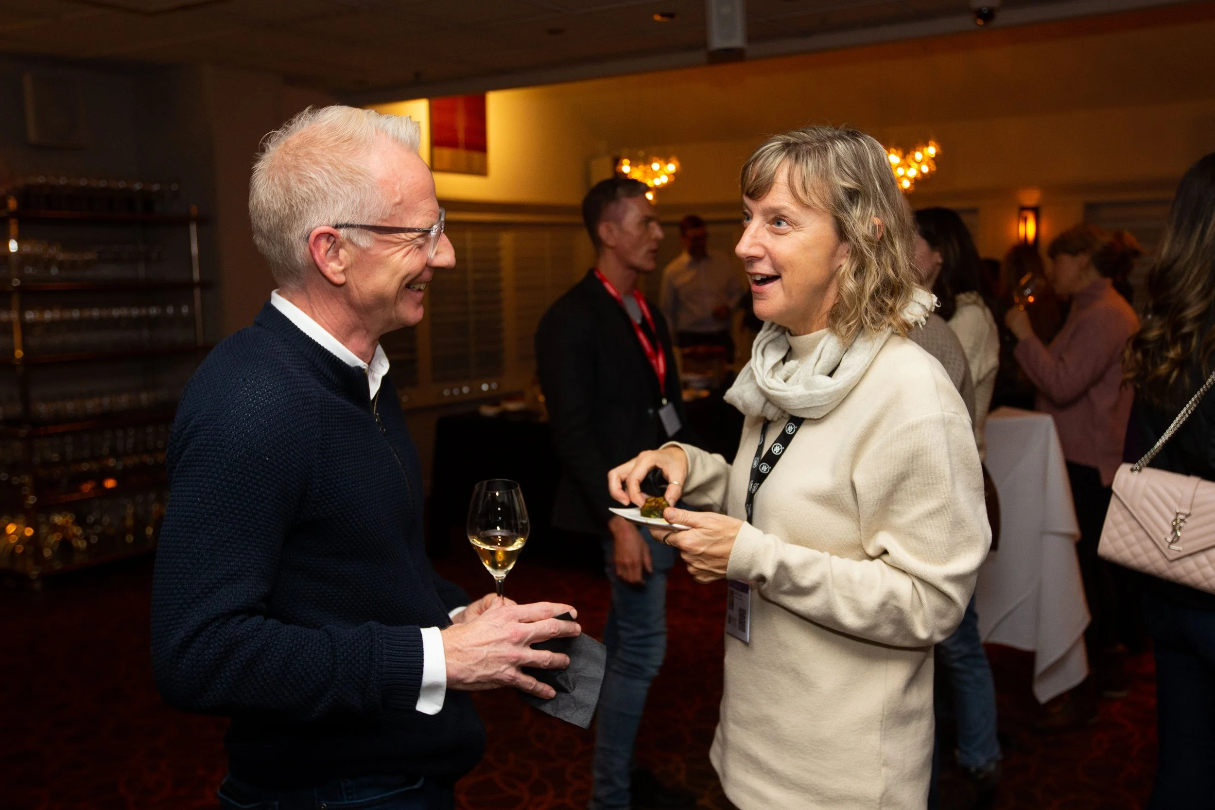 People networking at a social event indoors, holding drinks and food, engaging in conversation.