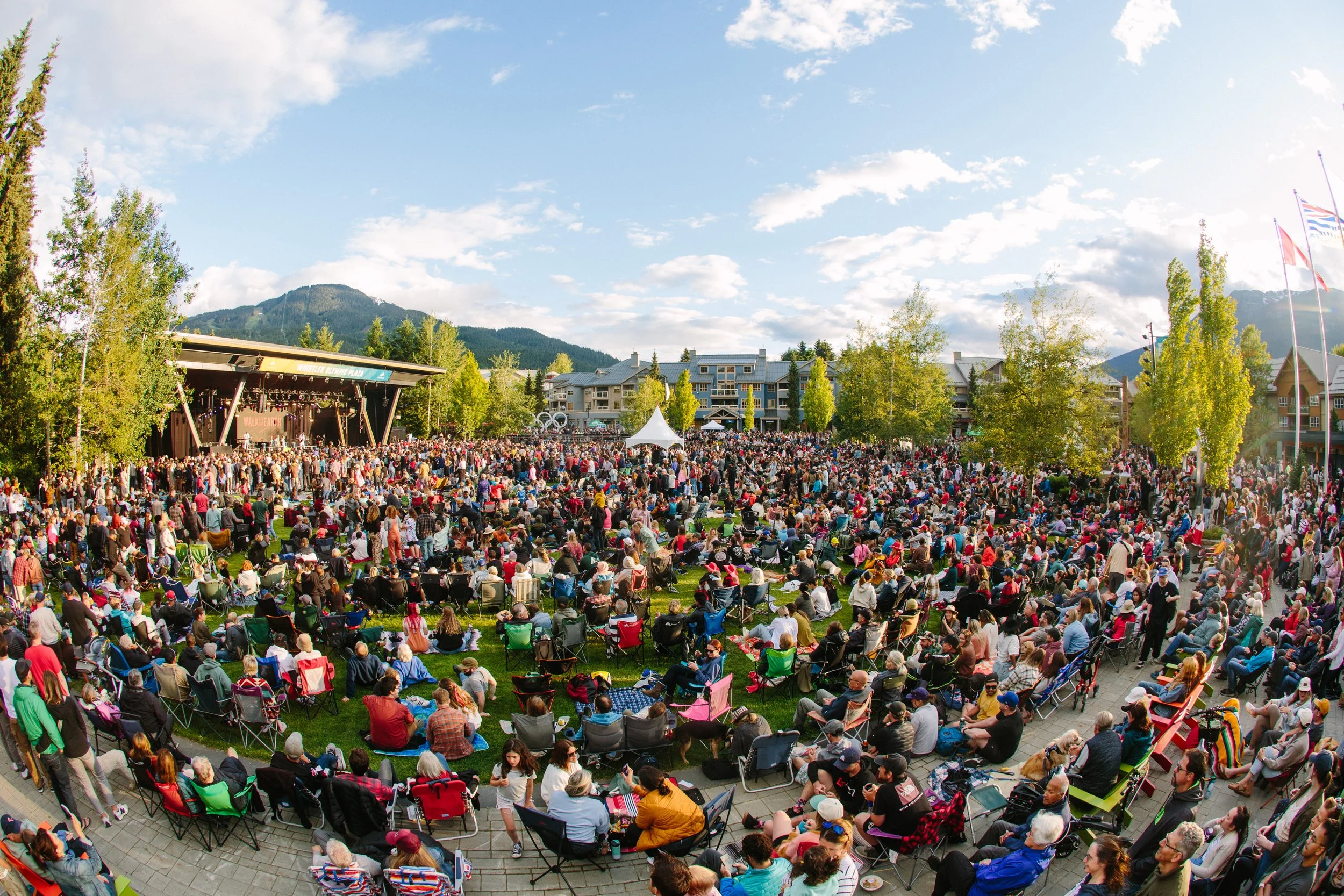 Large outdoor concert with a densely packed audience seated on grass and chairs, stage on the left, trees and mountains in the background, partly cloudy sky.