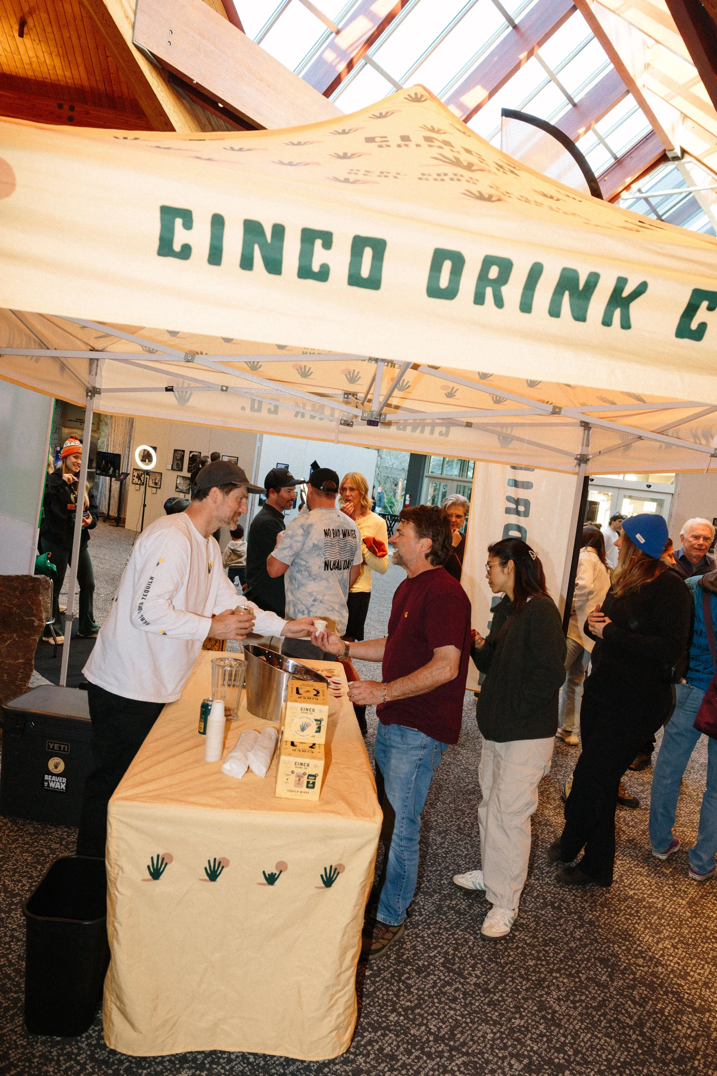 People gathered at a booth with a cream-colored tablecloth bearing the words "CINCO DRINK C" and illustrations of hands. A man is handing a cup to another person, with others in line or socializing nearby inside a building with glass ceiling and artw