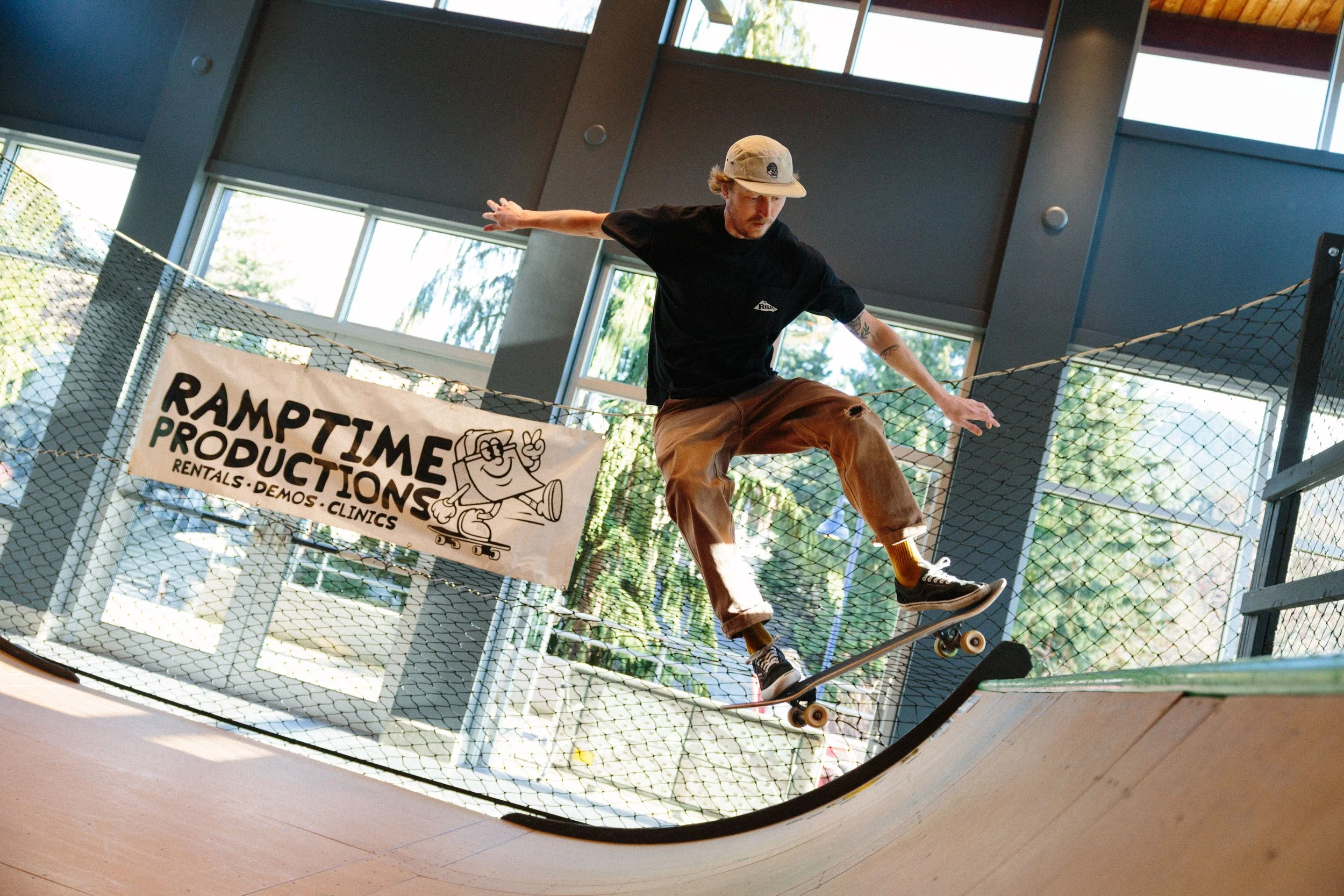 A young man skateboarding inside a skate park, balancing on a ledge with arms outstretched, wearing a tan baseball cap, a black t-shirt, brown pants, and black sneakers, with a fence and large windows in the background.