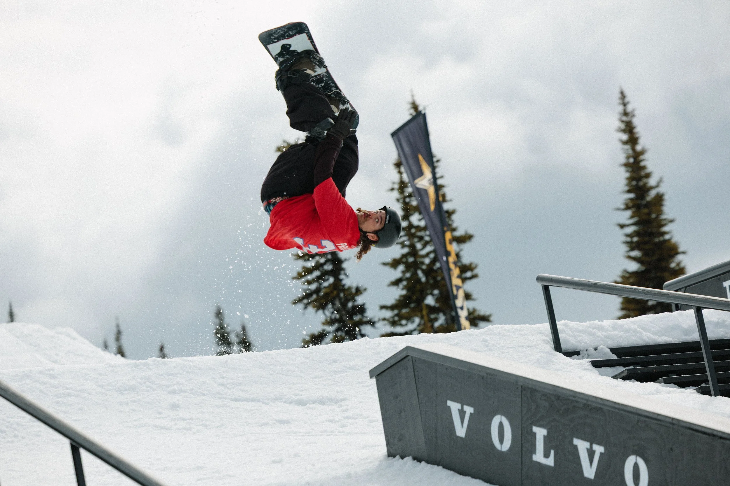 A snowboarder in a red jacket and black helmet executing a flip in mid-air over snow with tall evergreen trees and a cloudy sky in the background.