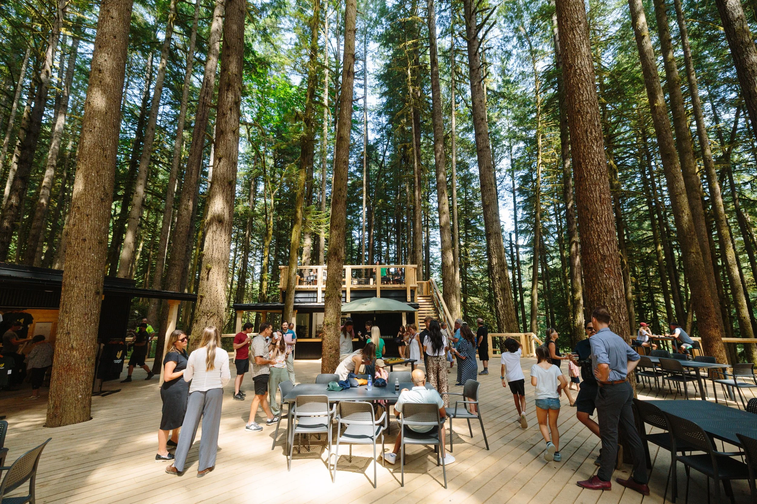 People gathered on a wooden deck in a forest, socializing and sitting around tables, with a treehouse structure in the background.