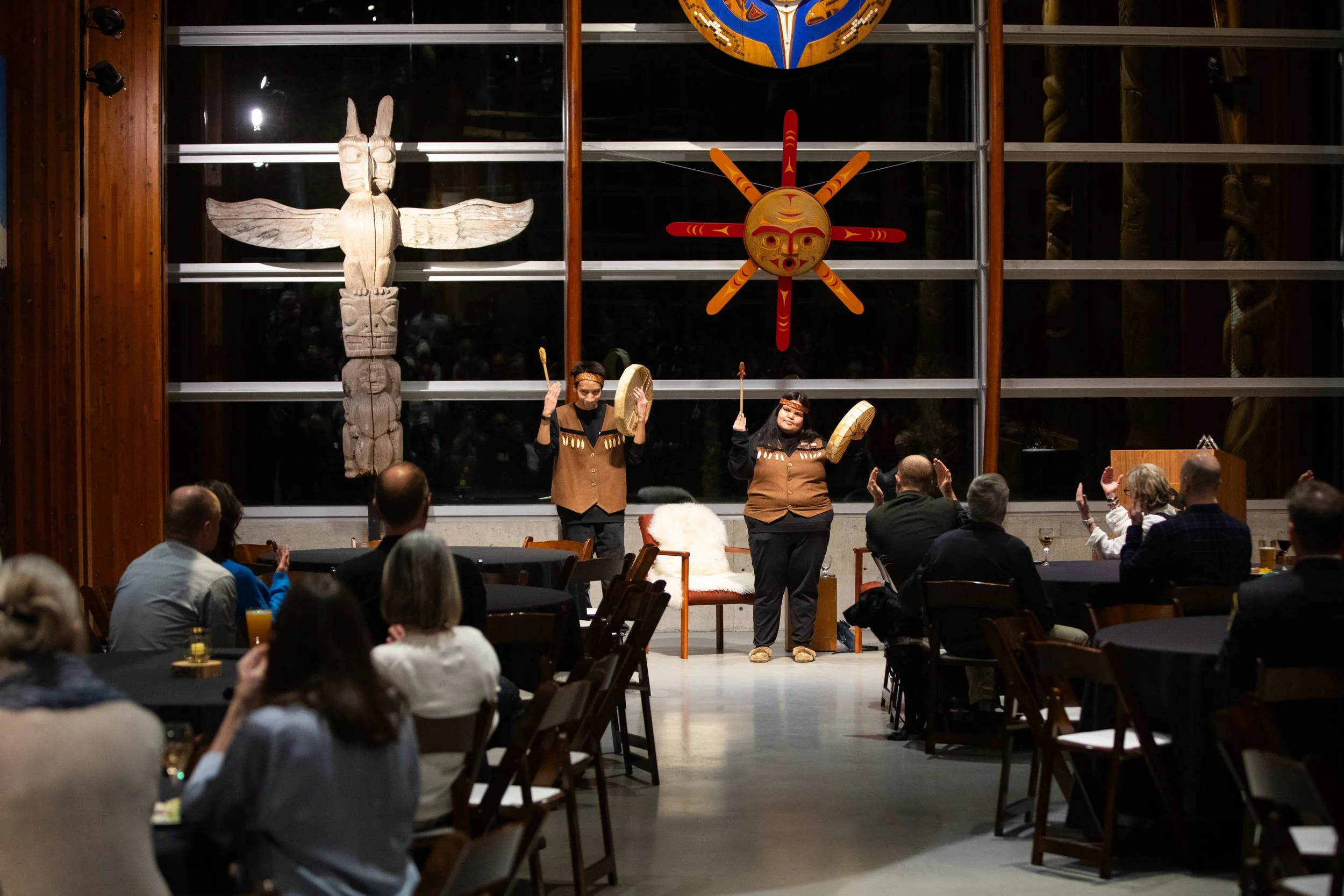 Two performers in traditional attire dance on stage with drums in front of a large wooden and textile artwork, while an audience watches seated at tables in a dimly lit room.