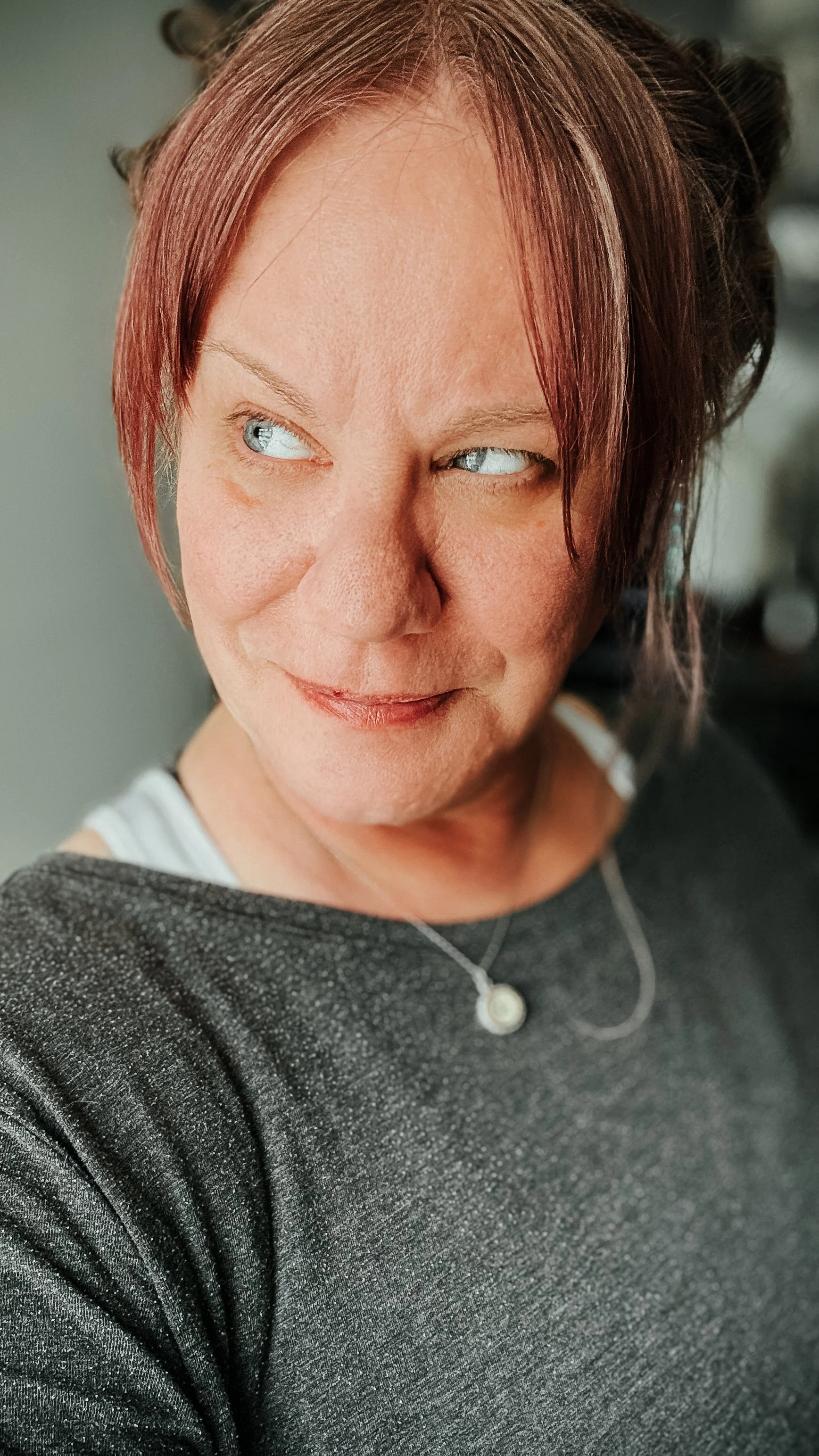Close-up of a woman with short reddish hair, blue eyes, wearing a dark gray top and a necklace, looking slightly to the side with a subtle smile.