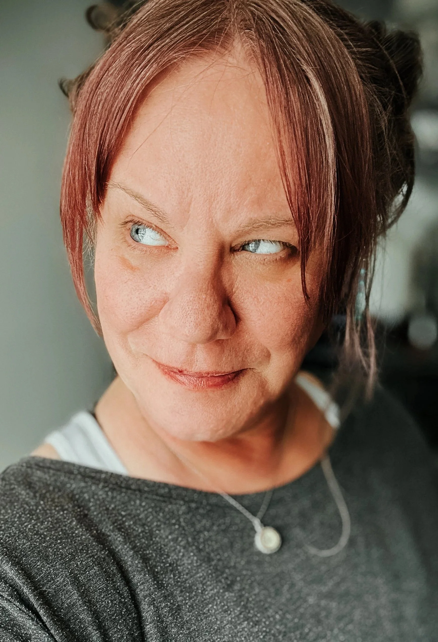 Close-up of a woman with short, reddish-brown hair styled in a messy bun, looking to her right, smiling softly. She has blue eyes, fair skin, and is wearing a black, glittery top with a white shirt underneath, and a silver necklace.