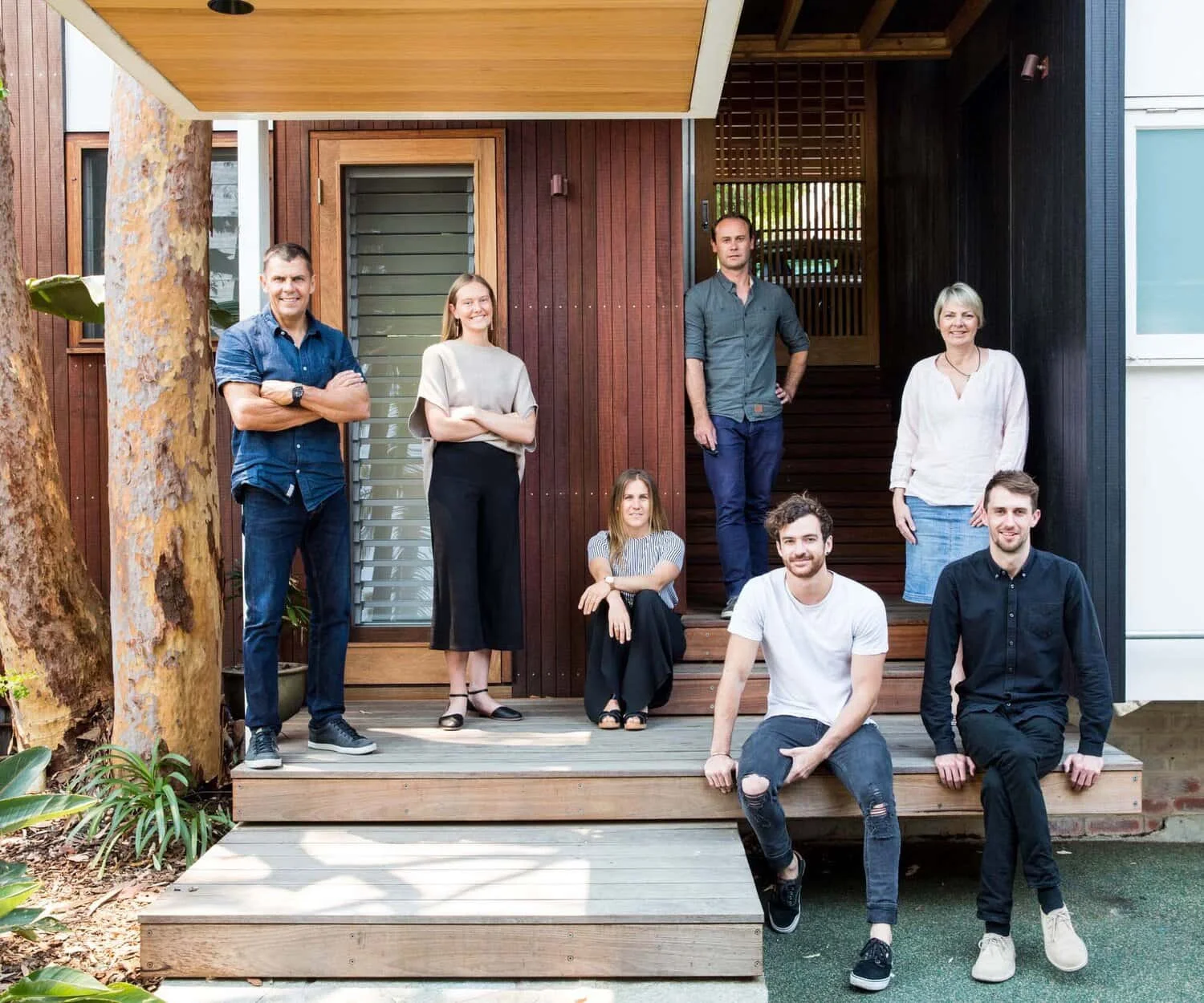 Team of seven professionals posing on wooden steps in front of a modern office building with a warm wood exterior and lush greenery.