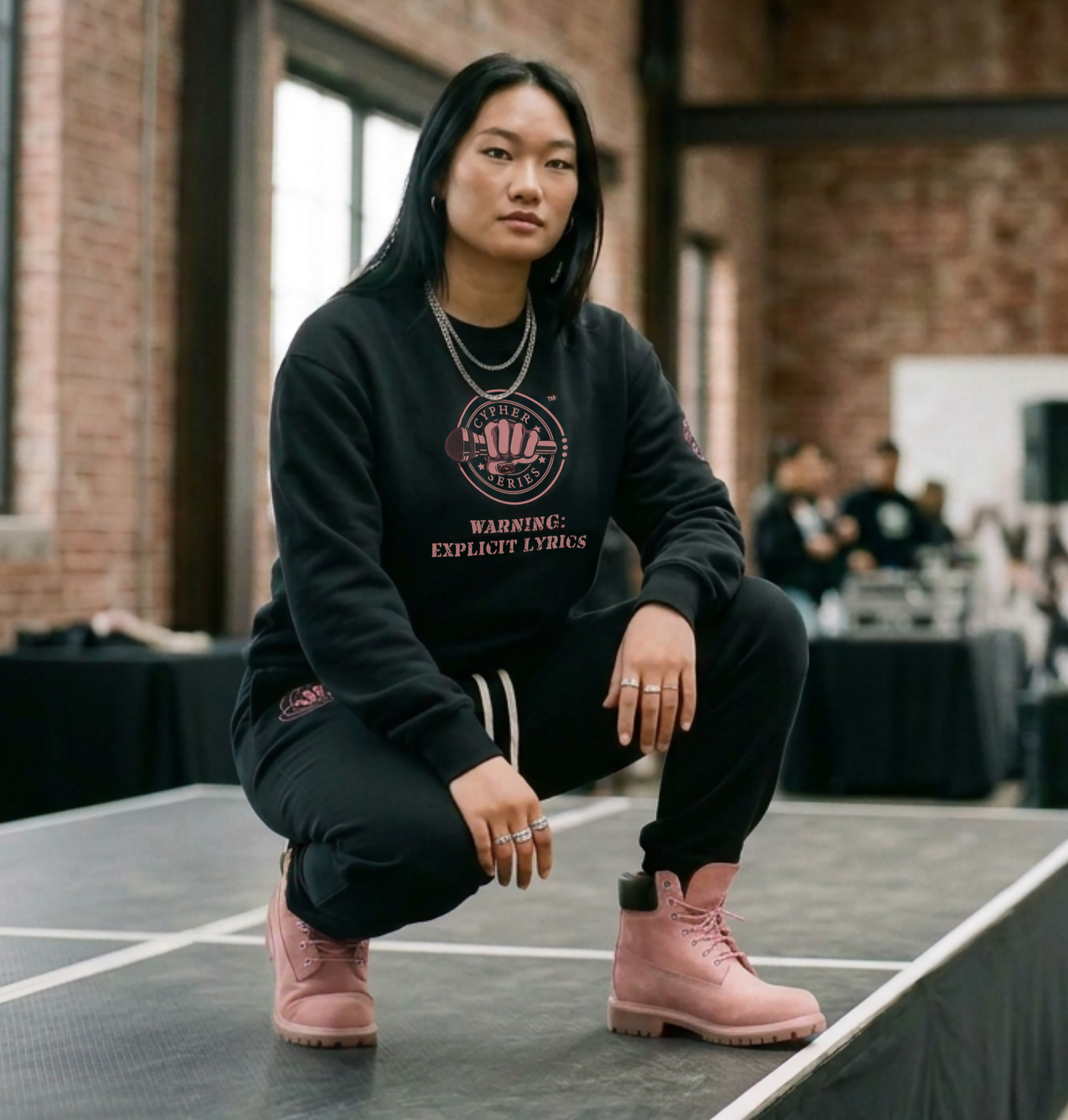 A woman with curly black hair wearing a black hoodie with a graphic design and black pants standing on railway tracks in a forested area.