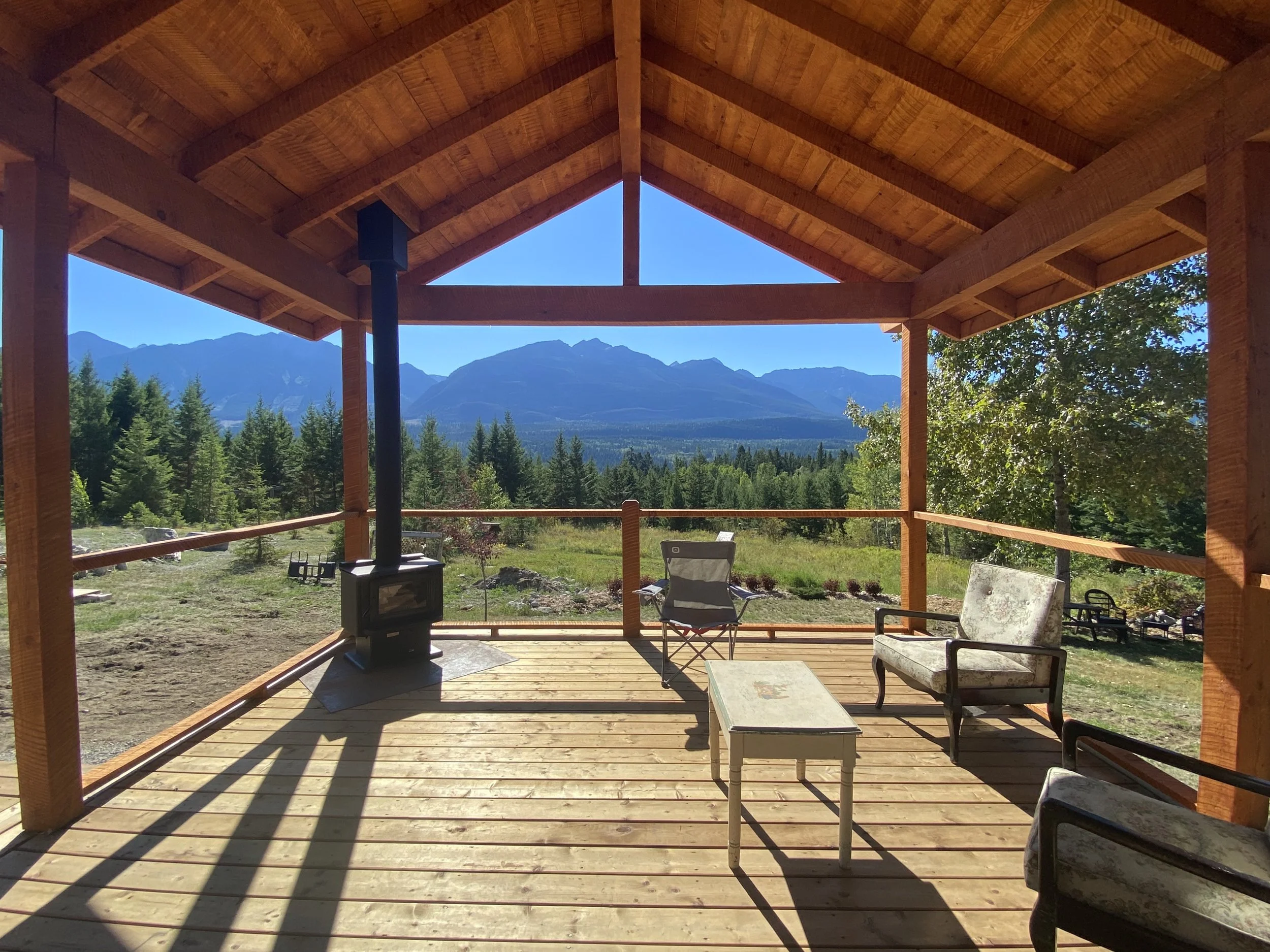Wooden porch with outdoor furniture including chairs, a small table, a wood stove, overlooking green trees and mountain range in the distance under a clear blue sky.