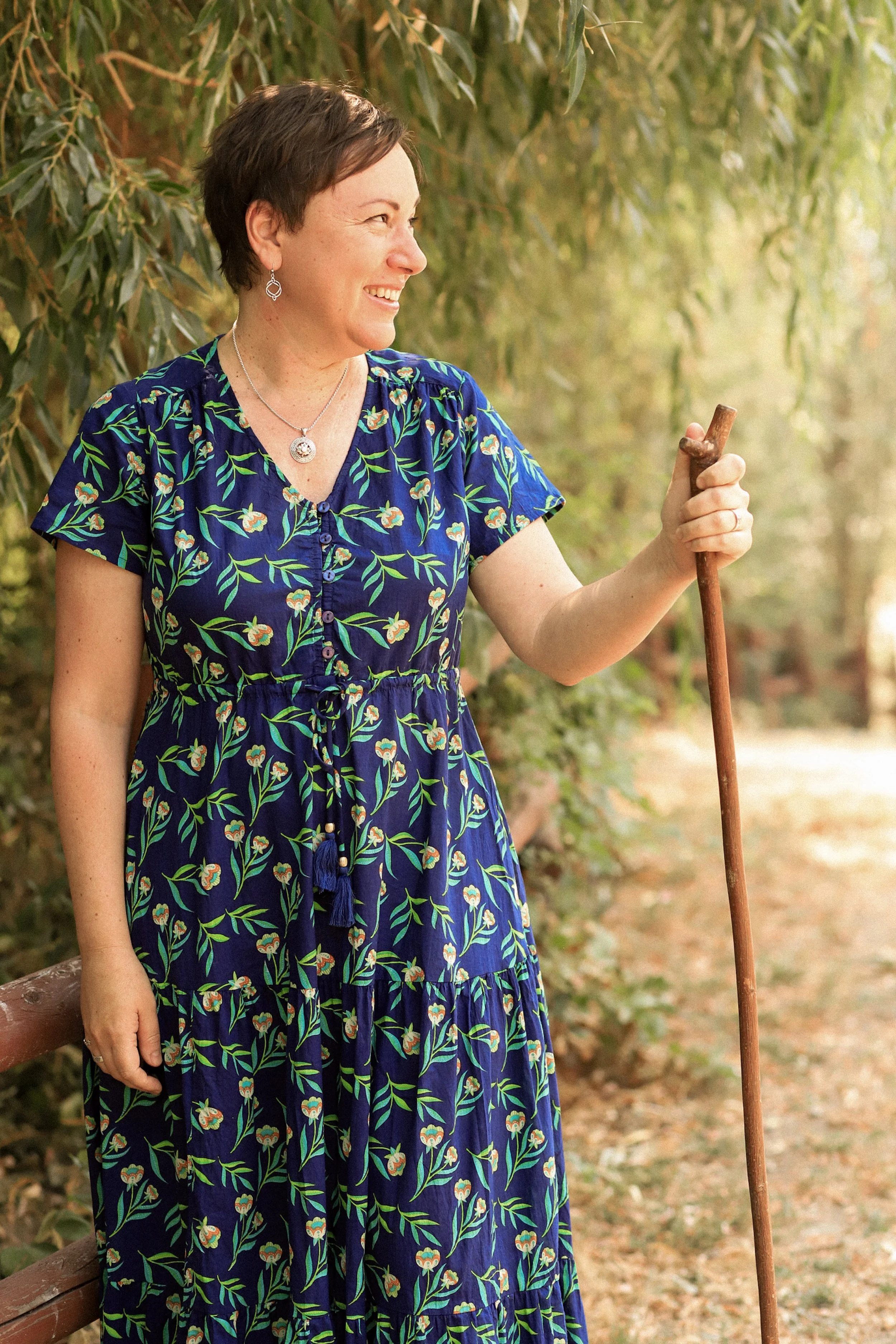 A woman with short brown hair, wearing a blue floral dress and jewelry, holding a walking stick, outdoors surrounded by trees.
