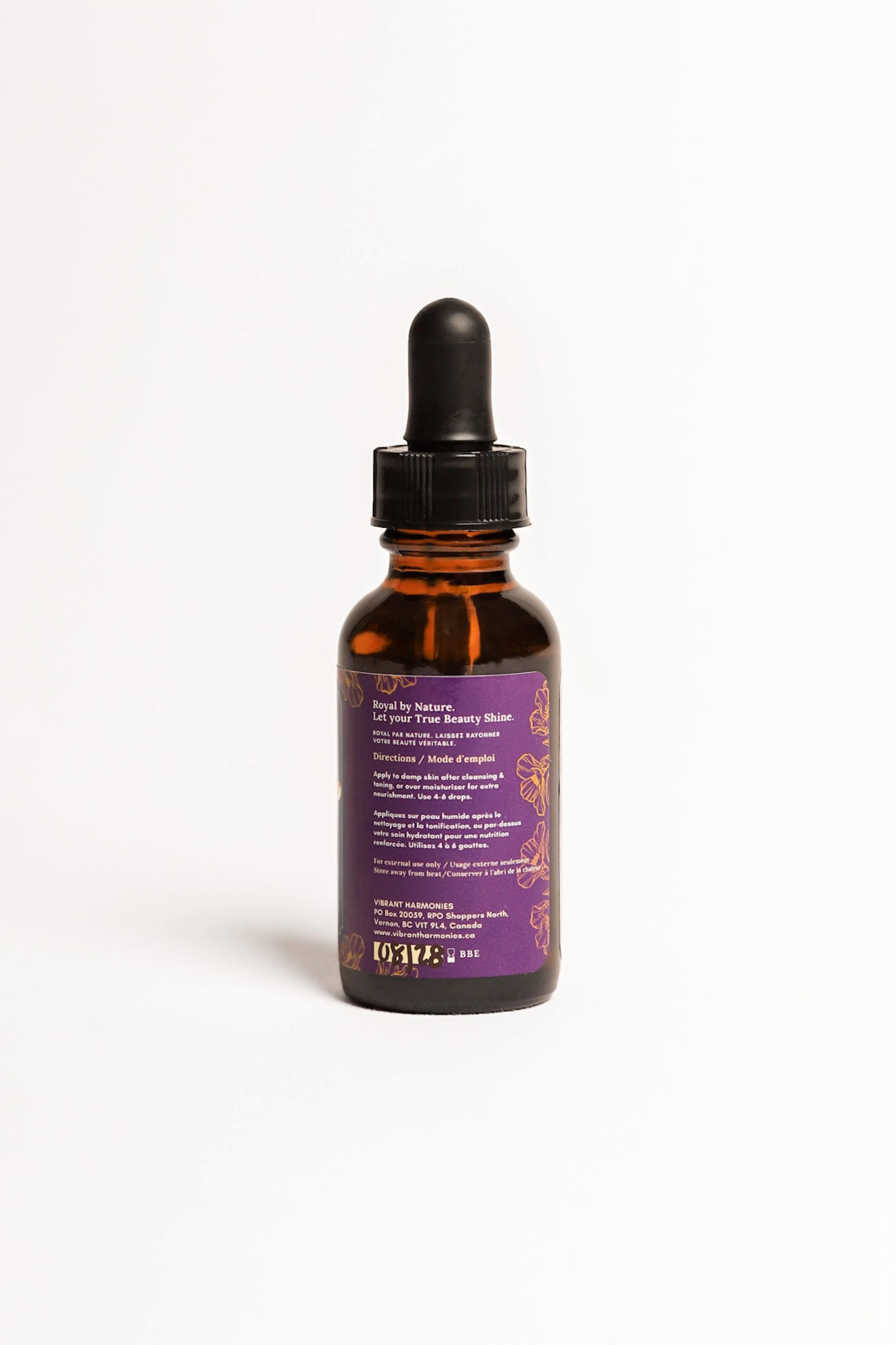 A small amber glass bottle with a black dropper cap. The bottle has a purple label with gold and white text, including directions for use and address information.