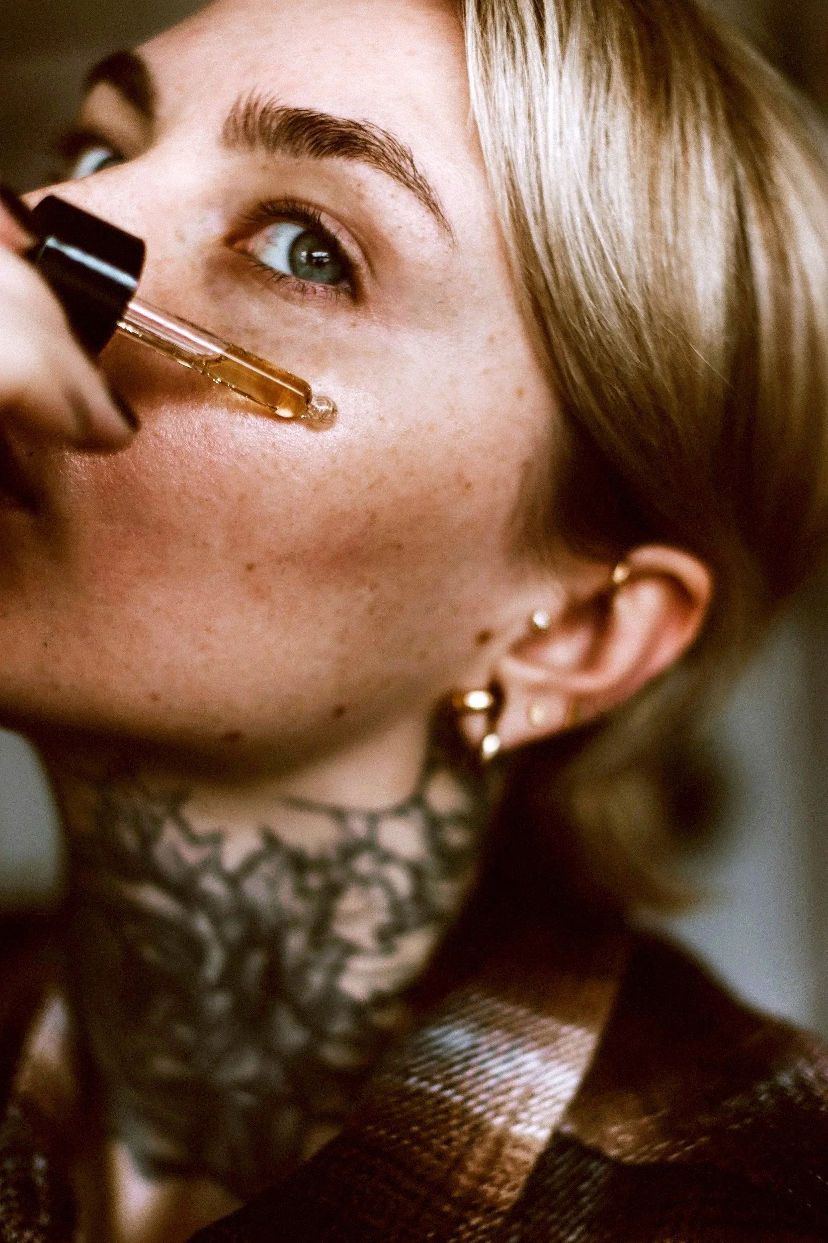 Close-up of a woman with blue eyes, light skin freckles, gold earrings, and long brown hair, applying lip gloss or similar product to her lips.